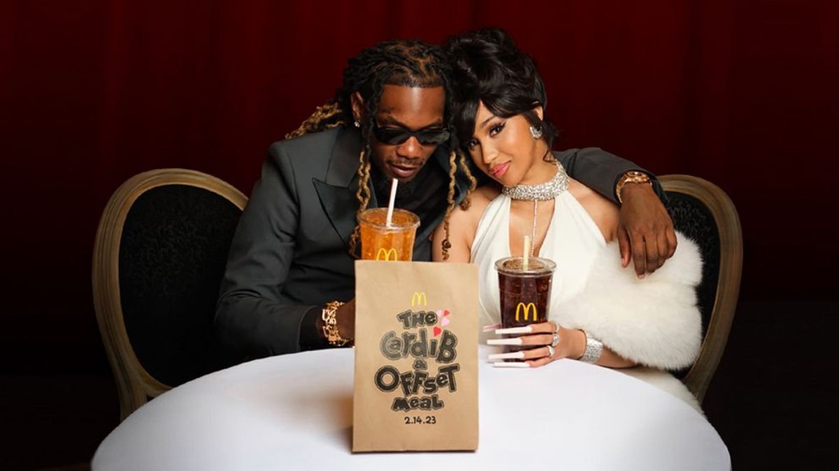 A man and woman sitting at a restaurant table with drinks, embracing, and smiling at the camera; the man is wearing sunglasses and a dark blazer, the woman has black hair styled in waves and is wearing a white dress with jewelry; there is a McDonald's bag on the table.