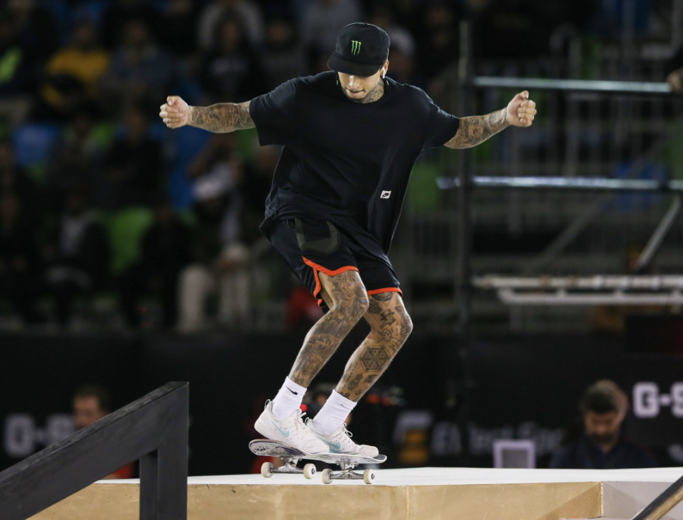 Skateboarder with tattoos wearing a black t-shirt, shorts, and a black cap with a Monster Energy logo performing a trick at a skate park with spectators in the background.
