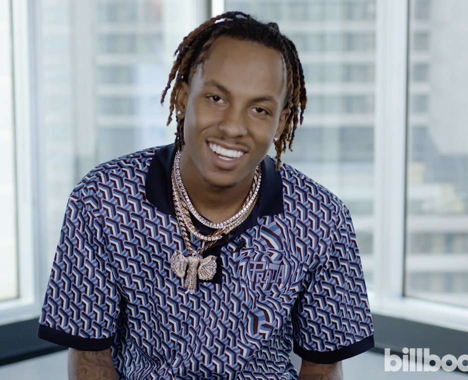 A man with medium-length dreadlocks, wearing diamond jewelry, smiling, in a patterned shirt, standing in front of large windows in a modern building.