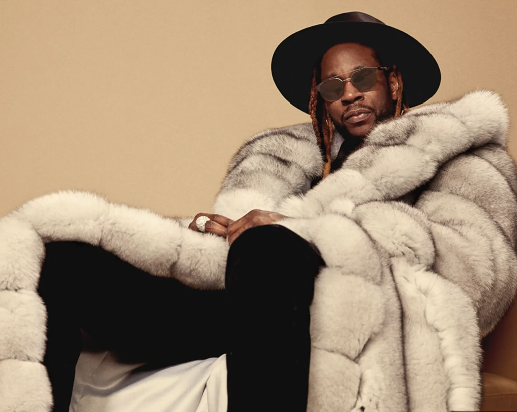 A man wearing sunglasses, a black wide-brim hat, and a luxurious fur coat, sitting against a beige background.