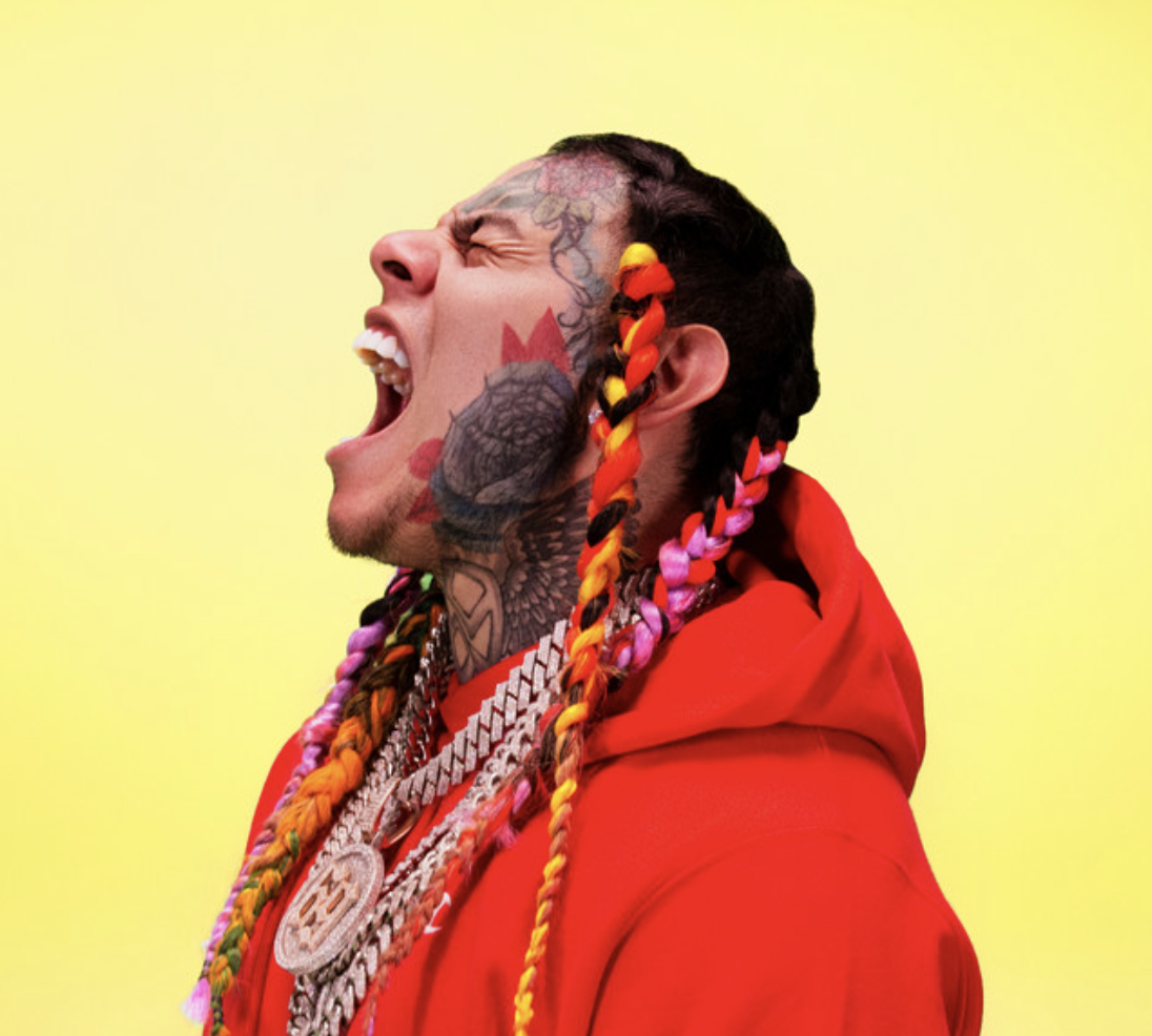 A man with colorful braided hair, tattoos on his face and neck, wearing a red hoodie and multiple chains, appears to be shouting or singing against a yellow background.