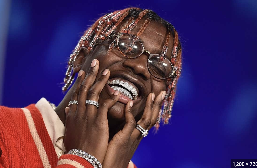 A smiling person with pink and blonde dreadlocks wearing glasses, diamond jewelry, a red and white varsity jacket, and a white shirt, posing against a blue background.