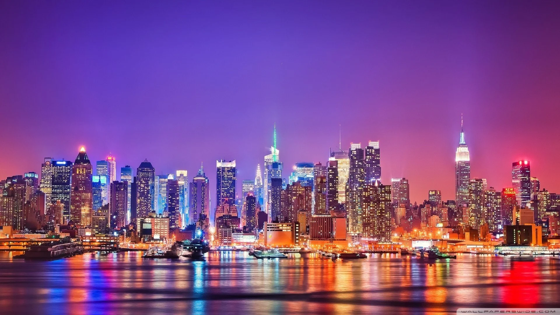 Nighttime city skyline of New York City with lit-up skyscrapers reflecting on a body of water in the foreground.