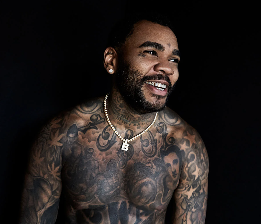 A shirtless man with numerous tattoos on his chest, arms, and neck, smiling and looking to his left against a black background.