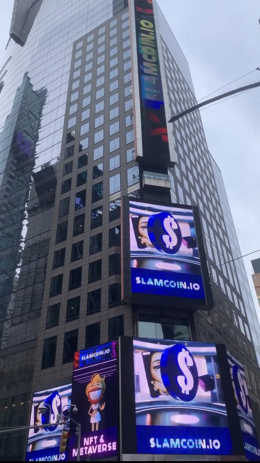 Digital billboards and screens on a tall building in Times Square advertising SLAMCOIN.IO, with bright colors, logos, Bitcoin symbols, and animated characters promoting NFTs and the metaverse.