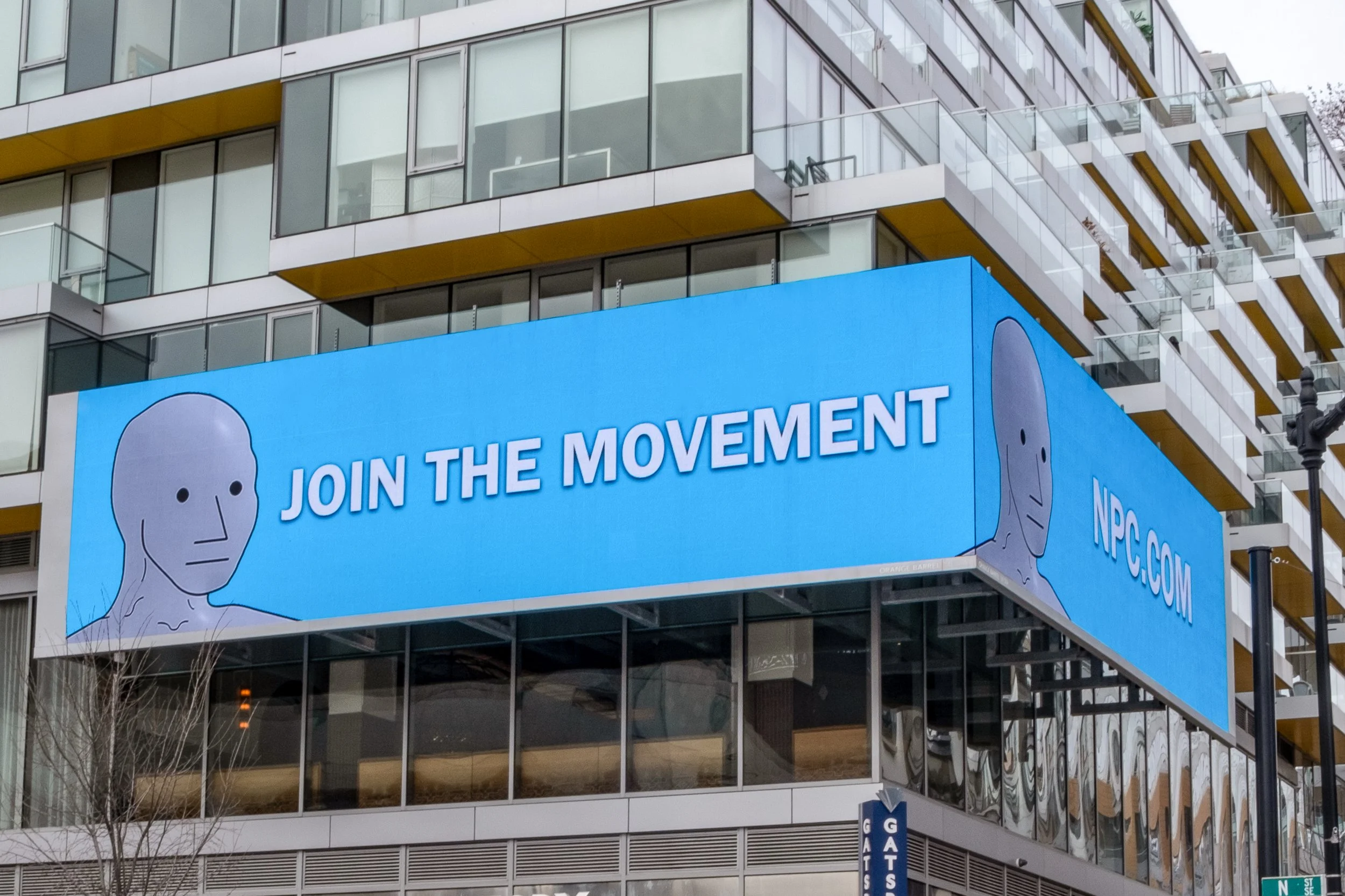 Large outdoor digital billboard on the side of a modern glass building displaying an advertisement for the NFT collection 'The Movement,' featuring a cartoonish face illustration and the text 'JOIN THE MOVEMENT'.