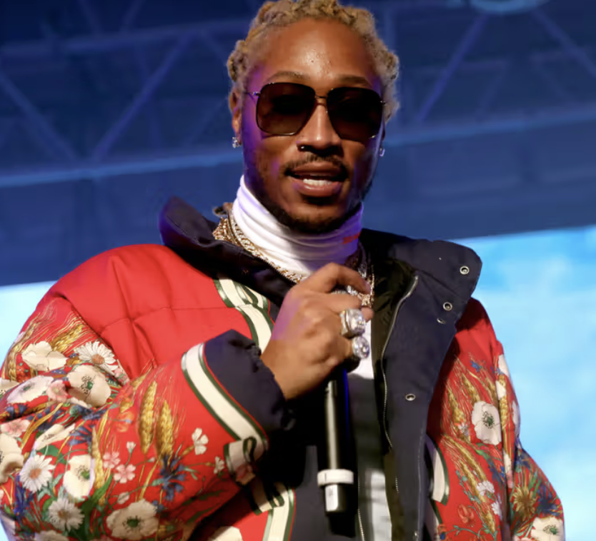 A man with blonde dreadlocks wearing sunglasses, a red patterned jacket, and jewelry, holding a microphone.