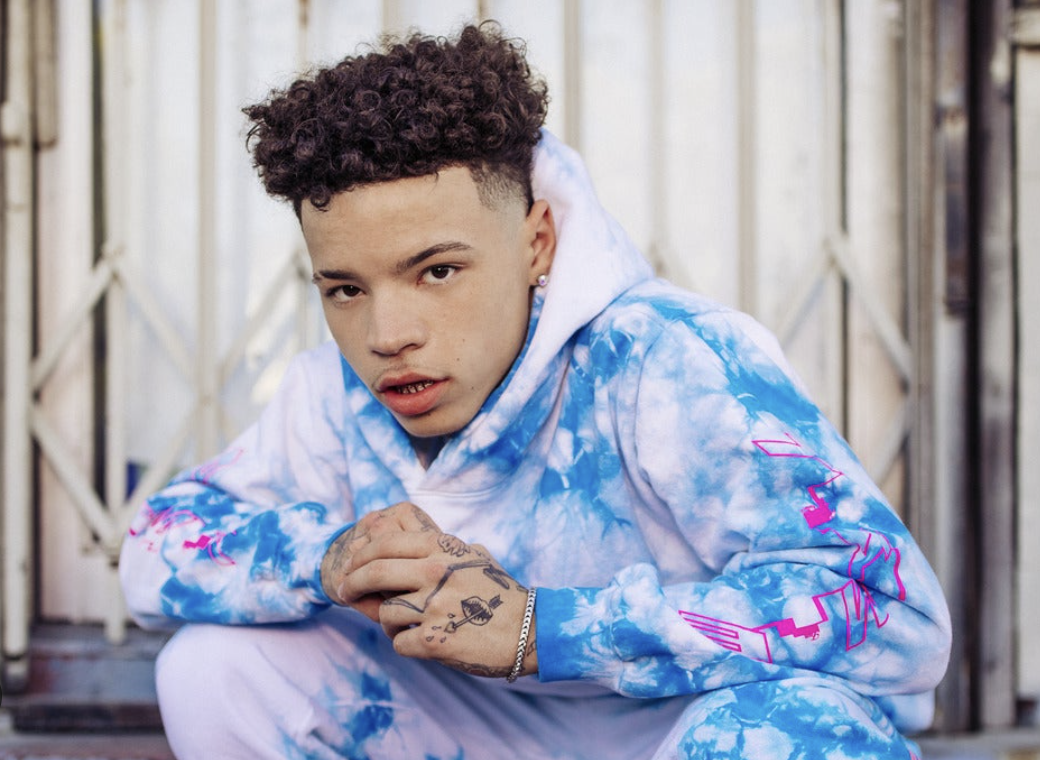 Young man with curly hair and tattoos on his hands, wearing a blue and white tie-dye hoodie, crouching outdoors with a serious expression.