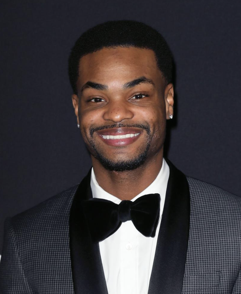 A young man in a tuxedo with a bow tie, smiling against a dark background.