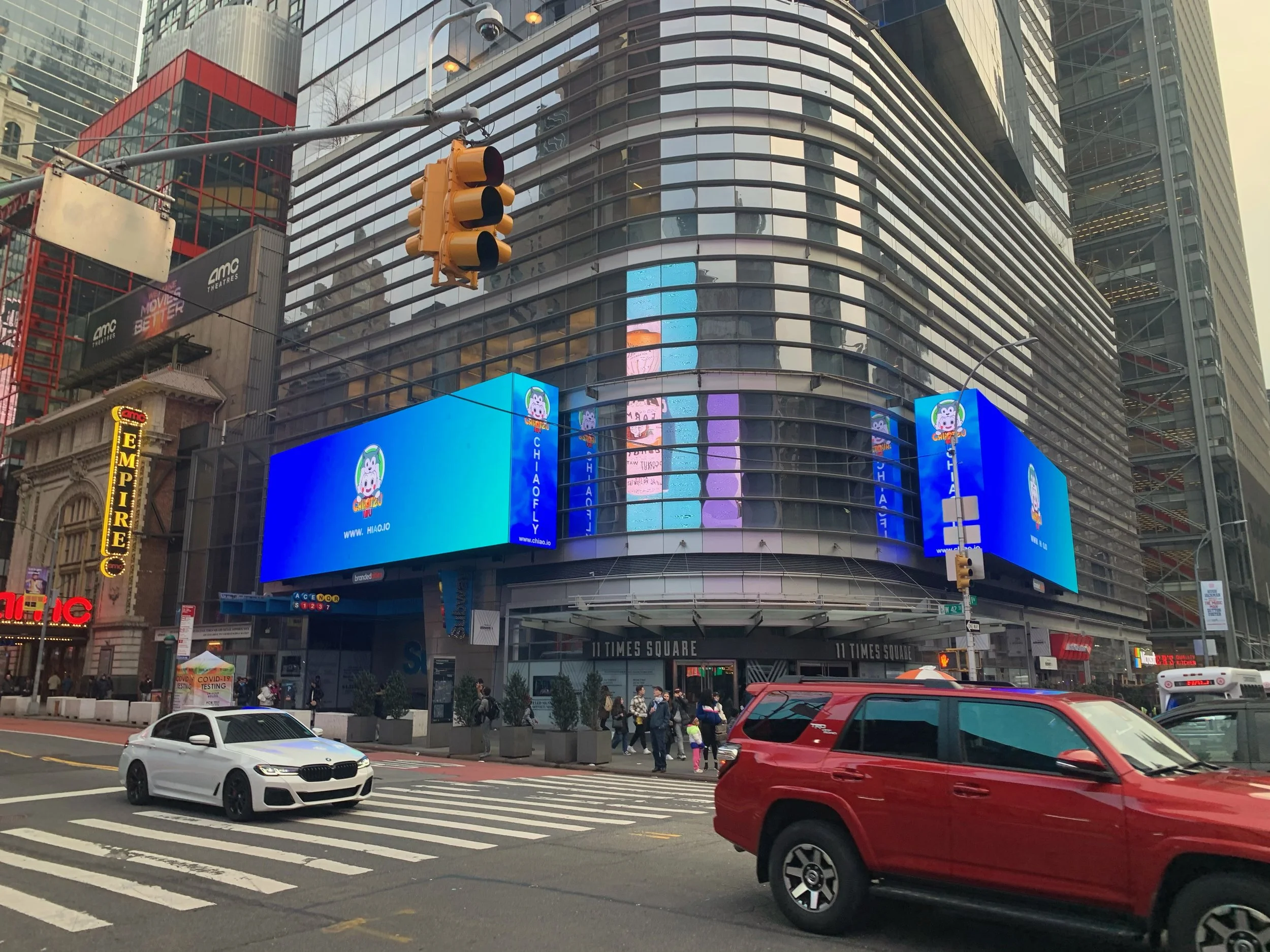 The image shows Times Square in New York City with tall buildings, digital billboards, and a busy city street with cars and pedestrians.