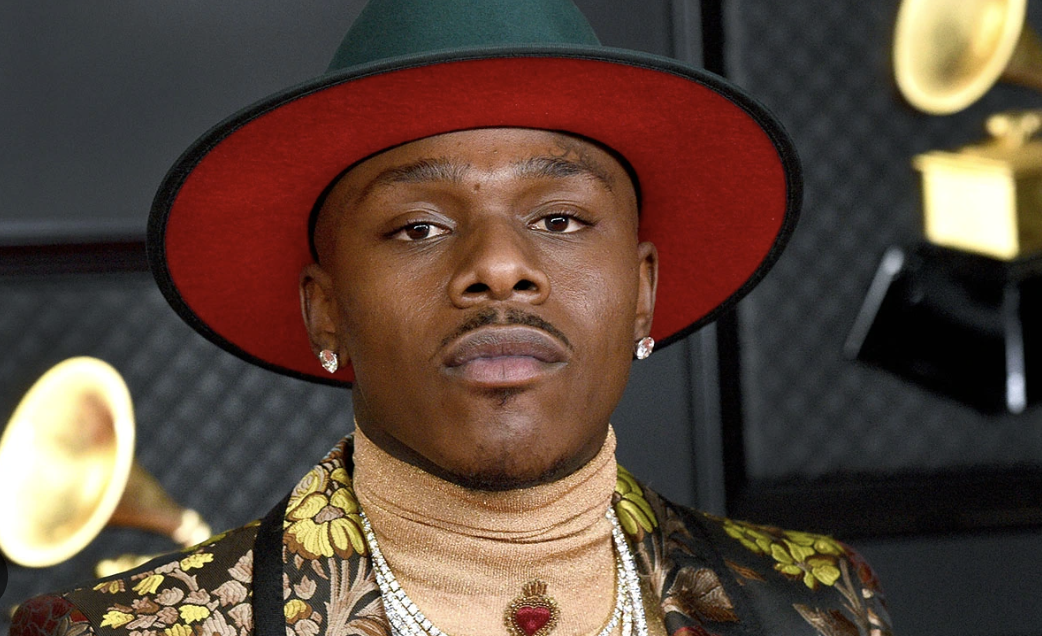 A man wearing a dark green and red wide-brimmed fedora hat, a floral-patterned blazer, a beige turtleneck, and jewelry, with a serious expression, in a room with audio equipment and yellow phonographs in the background.