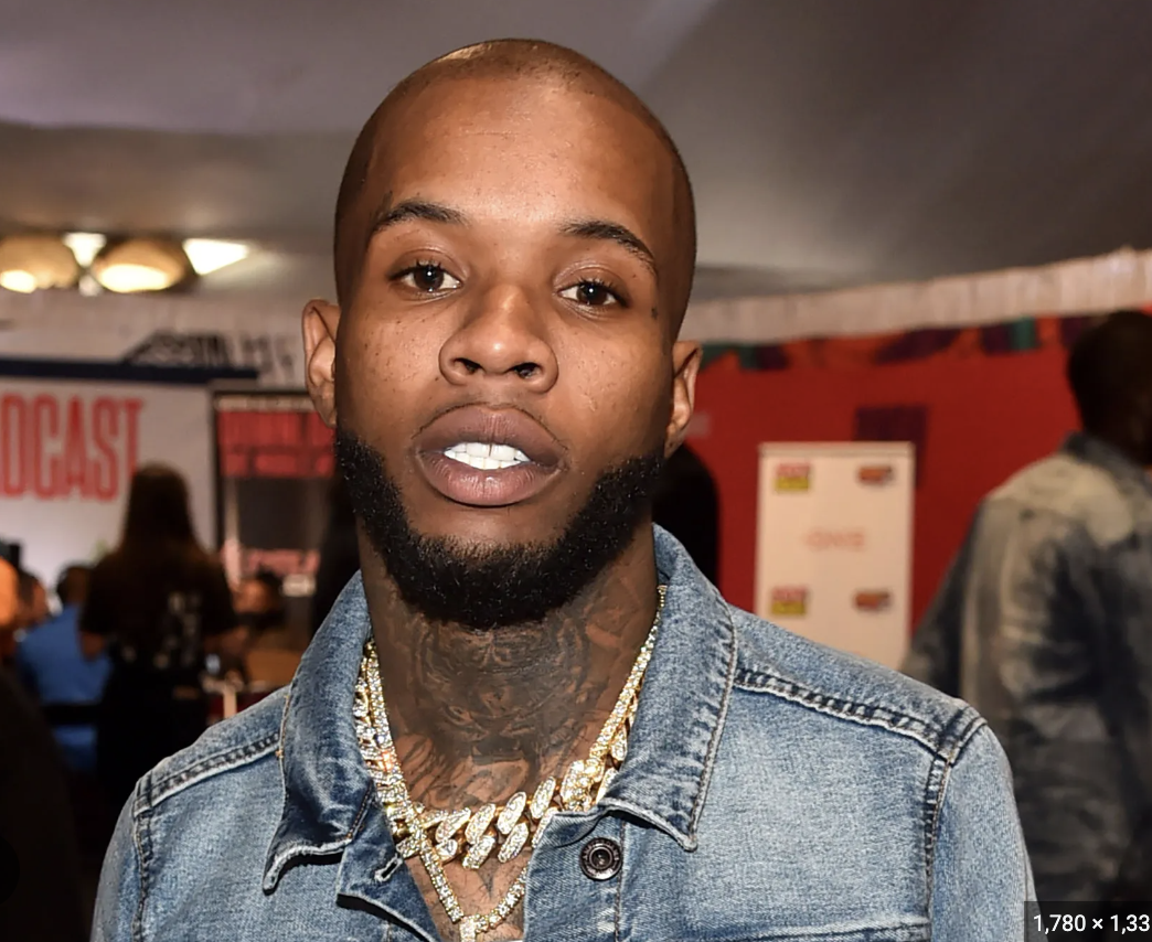 A young man with a shaved head and beard wearing a denim jacket and gold chains, standing indoors at a social event.
