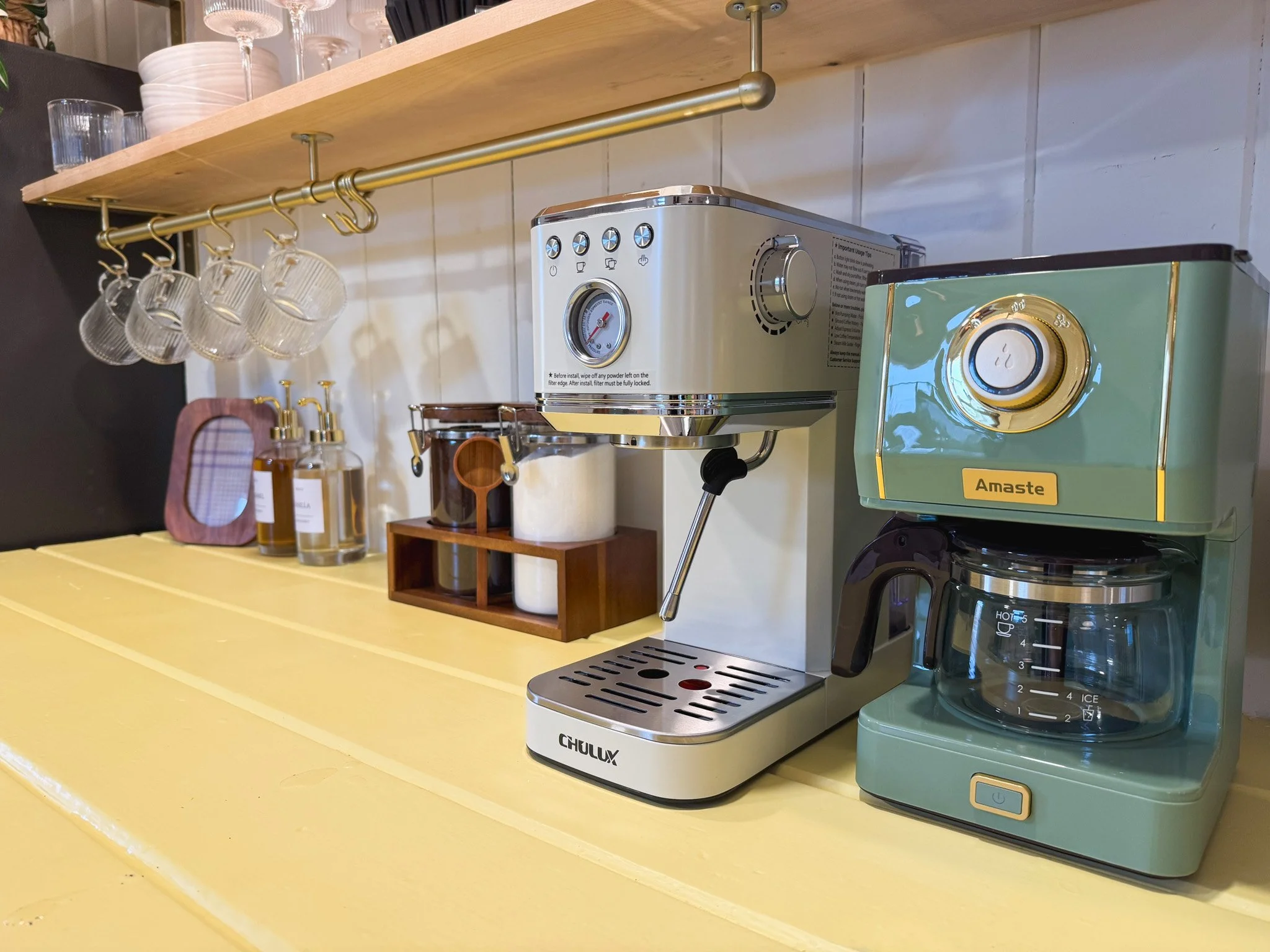 Coffee station with a silver espresso machine, a green coffee maker, hanging glass cups, and bottles of syrup on a yellow countertop.