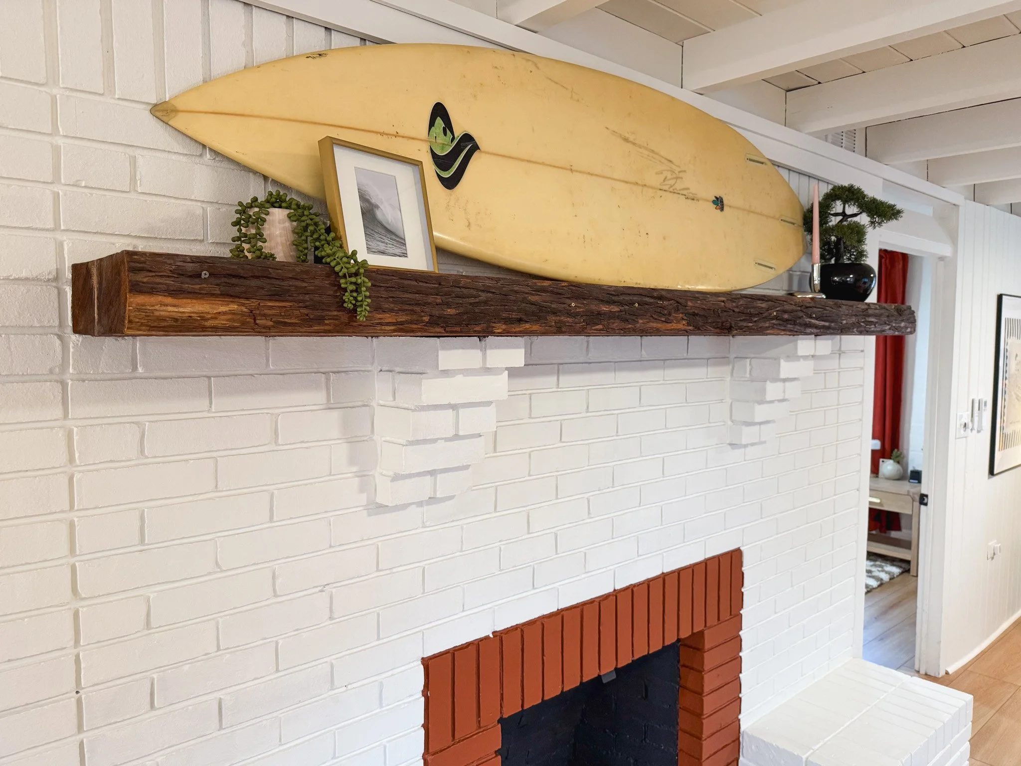 Interior of a cozy living room with a white painted brick fireplace, a rustic wooden mantel, a yellow surfboard, a framed black-and-white photo, a small potted plant, and various decorative items.