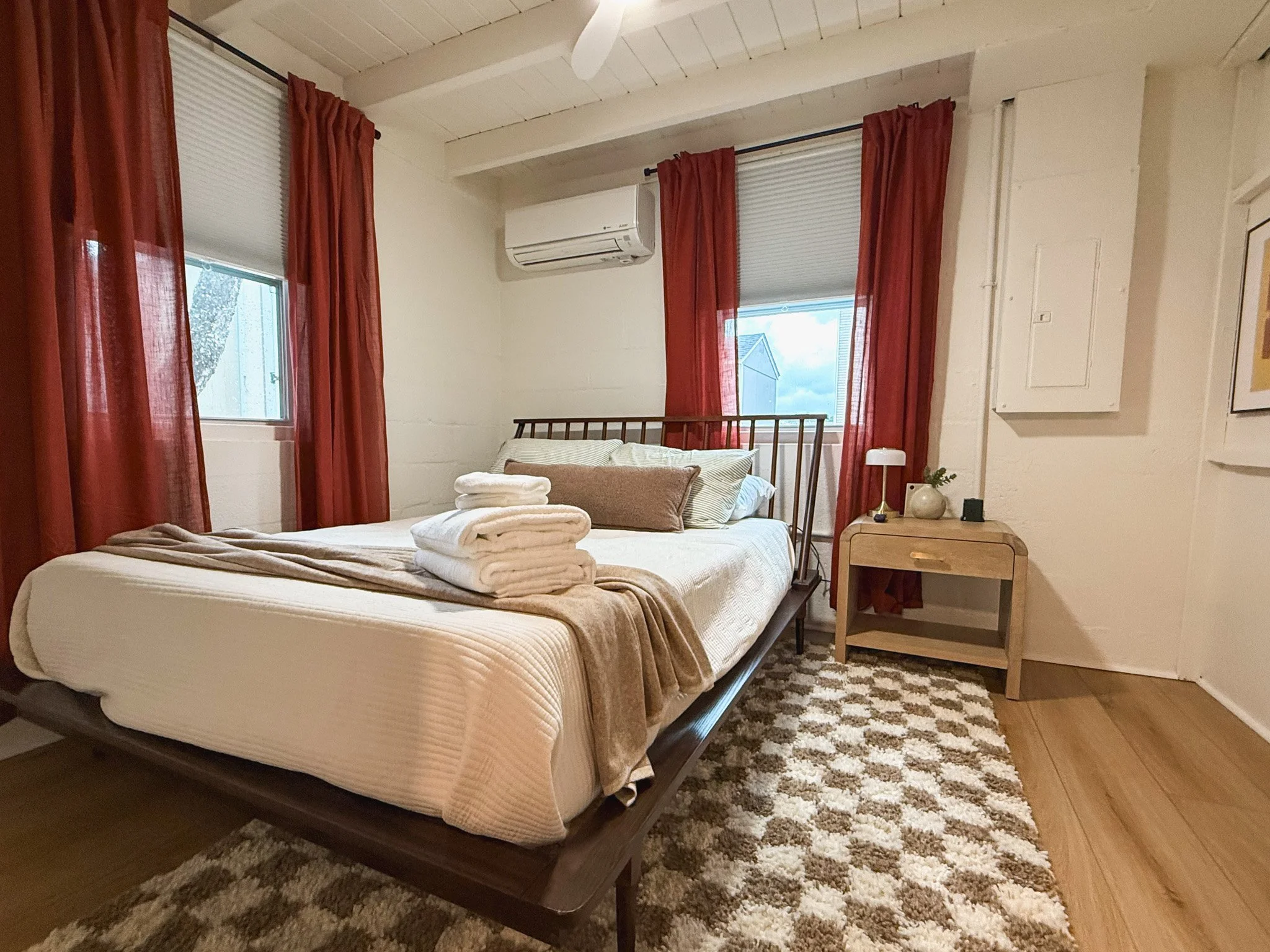 A cozy bedroom with a bed, red curtains, and a small wooden nightstand with a lamp and decor, situated on a plush patterned rug.