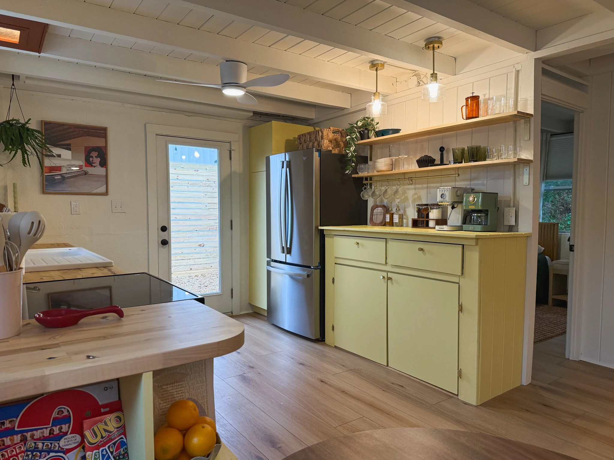 Kitchen with yellow cabinet, open shelves with dishes and glasses, stainless steel refrigerator, wooden floor, and door leading outside. Partially visible wooden table with lemons and a children's game in the foreground.