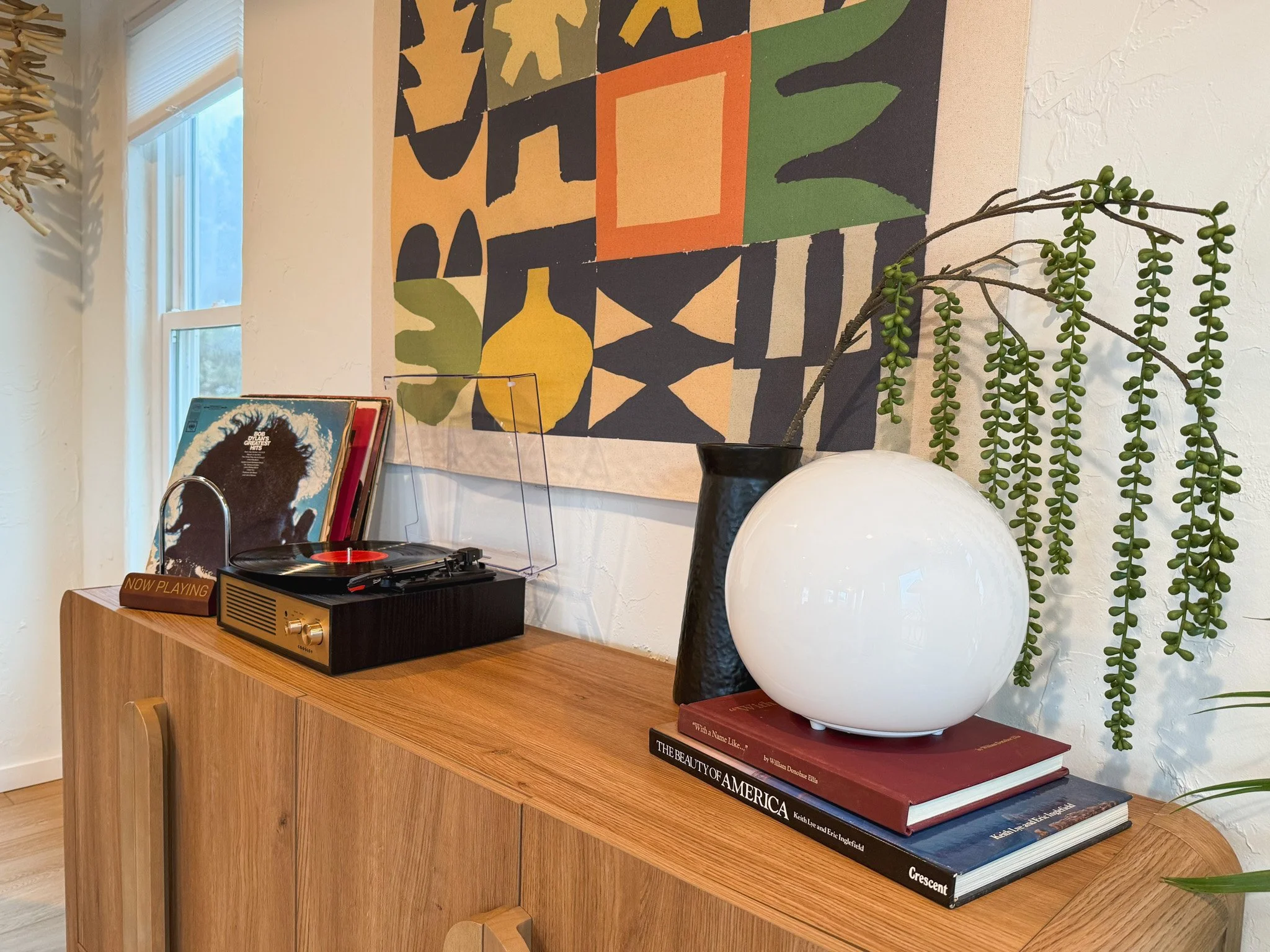 Decorative wooden sideboard topped with a vinyl record player, a stack of books, a white round vase, and a tall dark vase with a trailing green plant. Behind is a colorful abstract wall hanging and a window with natural light.