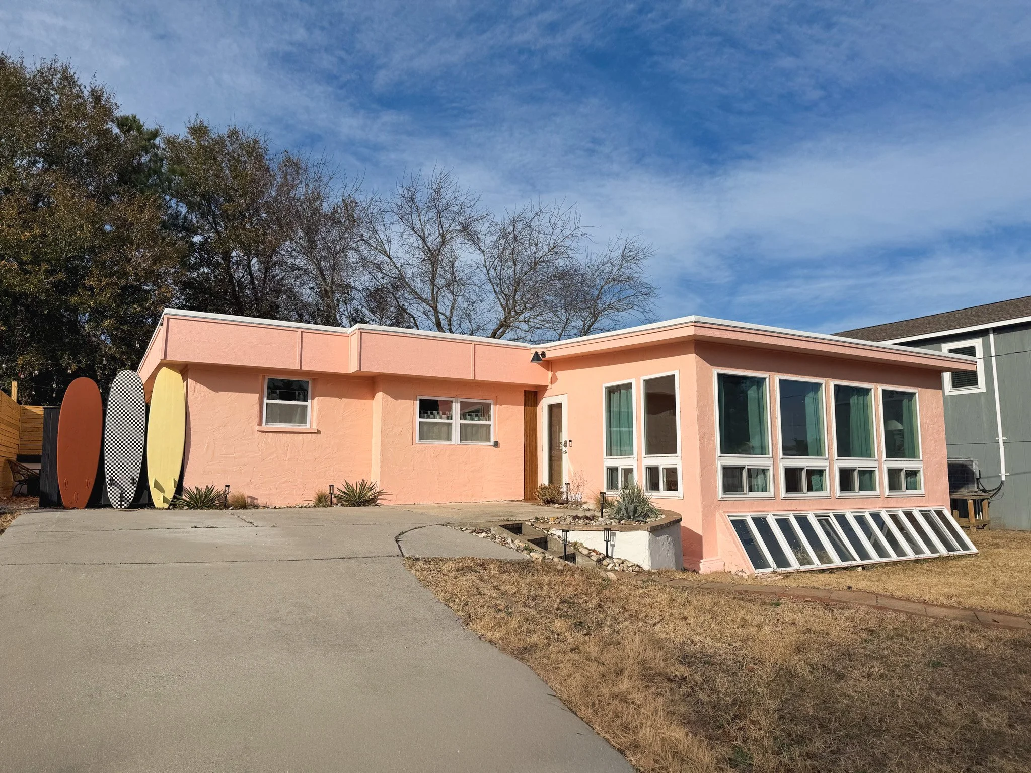 A pink single-story house with a curved driveway, large windows, and four surfboards of different colors leaning against the left side of the house under a blue sky with some clouds.