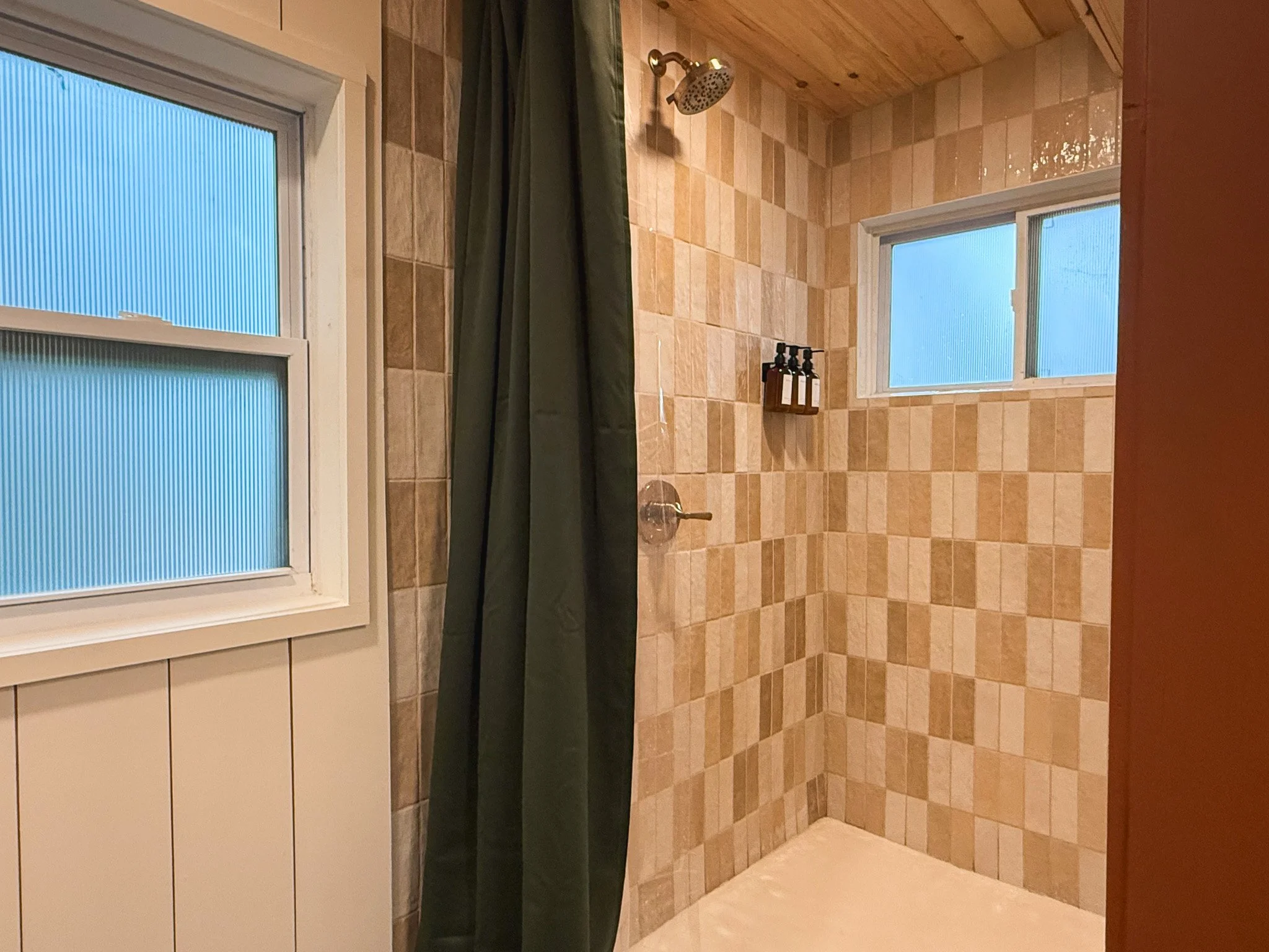 The image shows a shower area with beige and brown checkered tiles, a window with frosted glass, a dark green shower curtain, a silver showerhead, and three shampoo bottles on a small black rack.