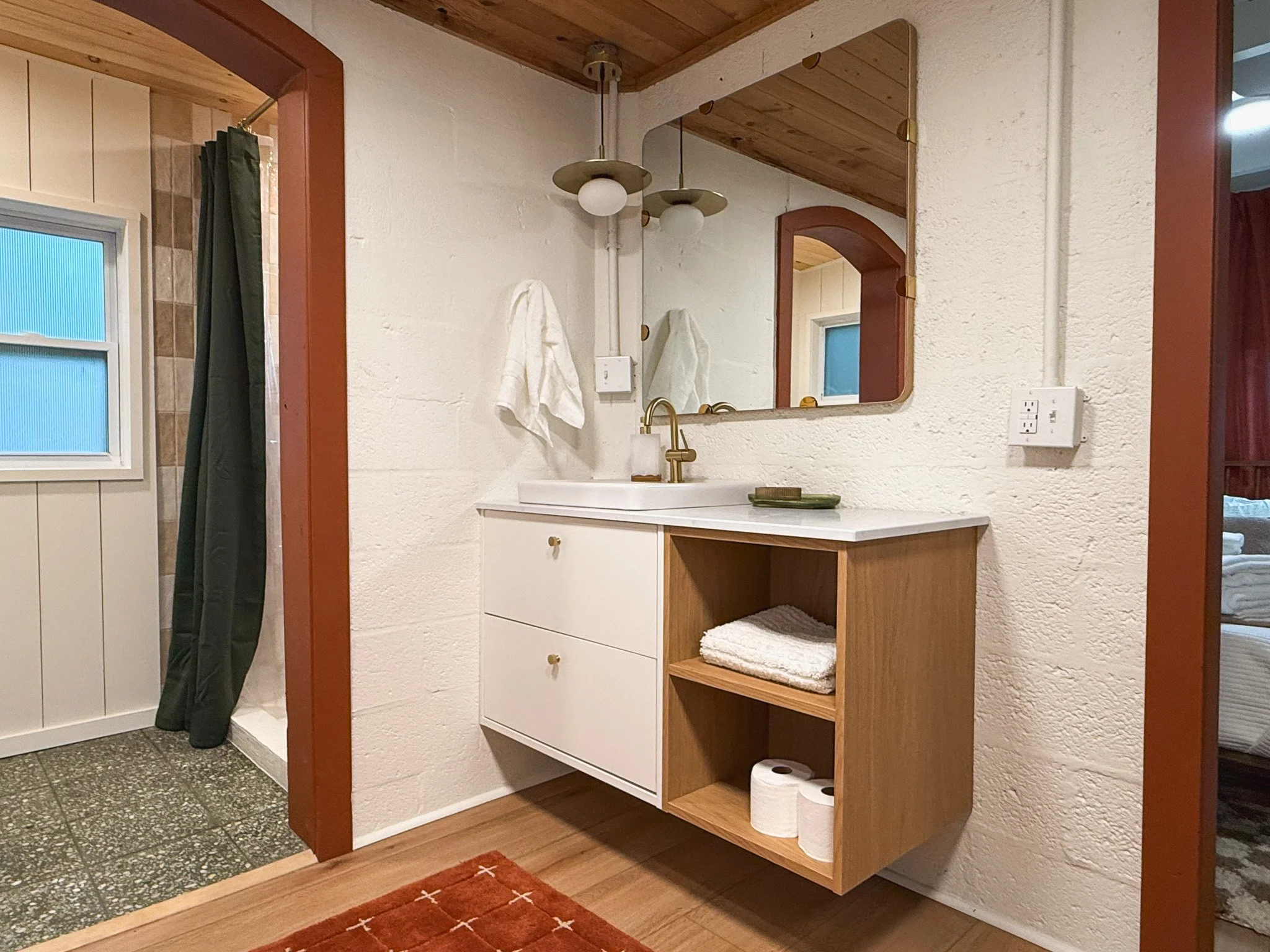 Bathroom with a white vanity, a mirror, and a gold faucet. Towels and toiletries on the vanity, open wooden shelving with towels and toilet paper. Adjacent shower area with dark green curtain and window, wooden ceiling, and textured white walls.