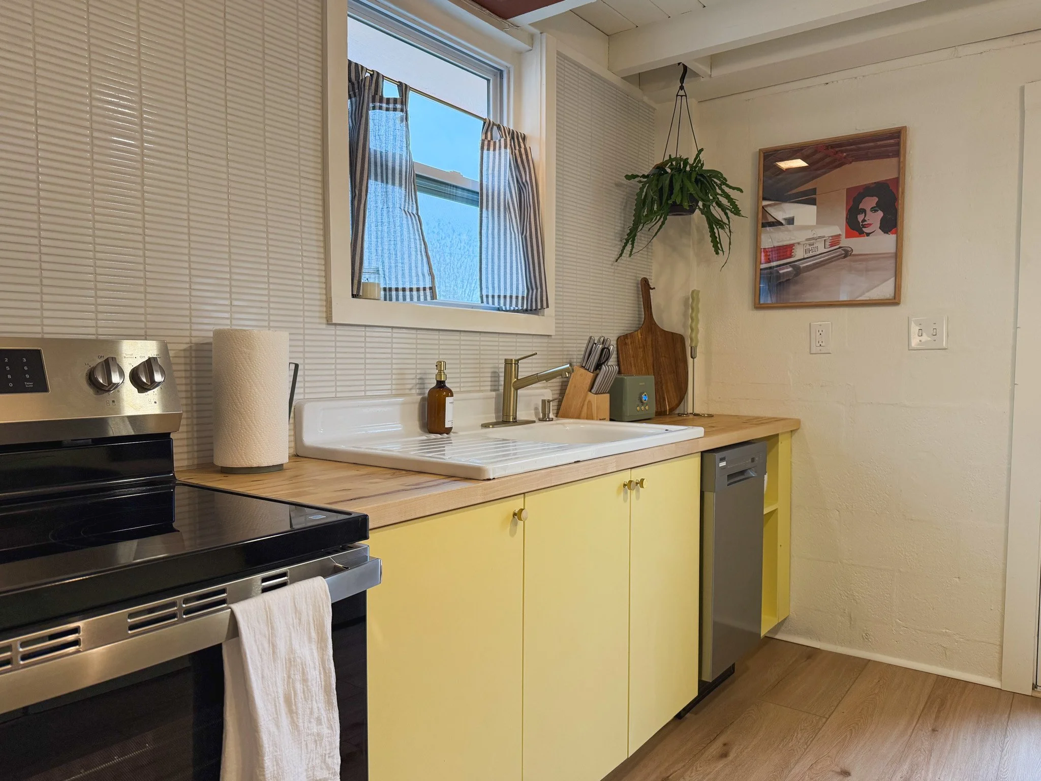 A cozy kitchen with a yellow lower cabinet, light wood countertop, and a black stove. There is a window with striped curtains above the sink, a paper towel roll, a soap dispenser, and some kitchen utensils. A hanging plant and a framed picture of a w