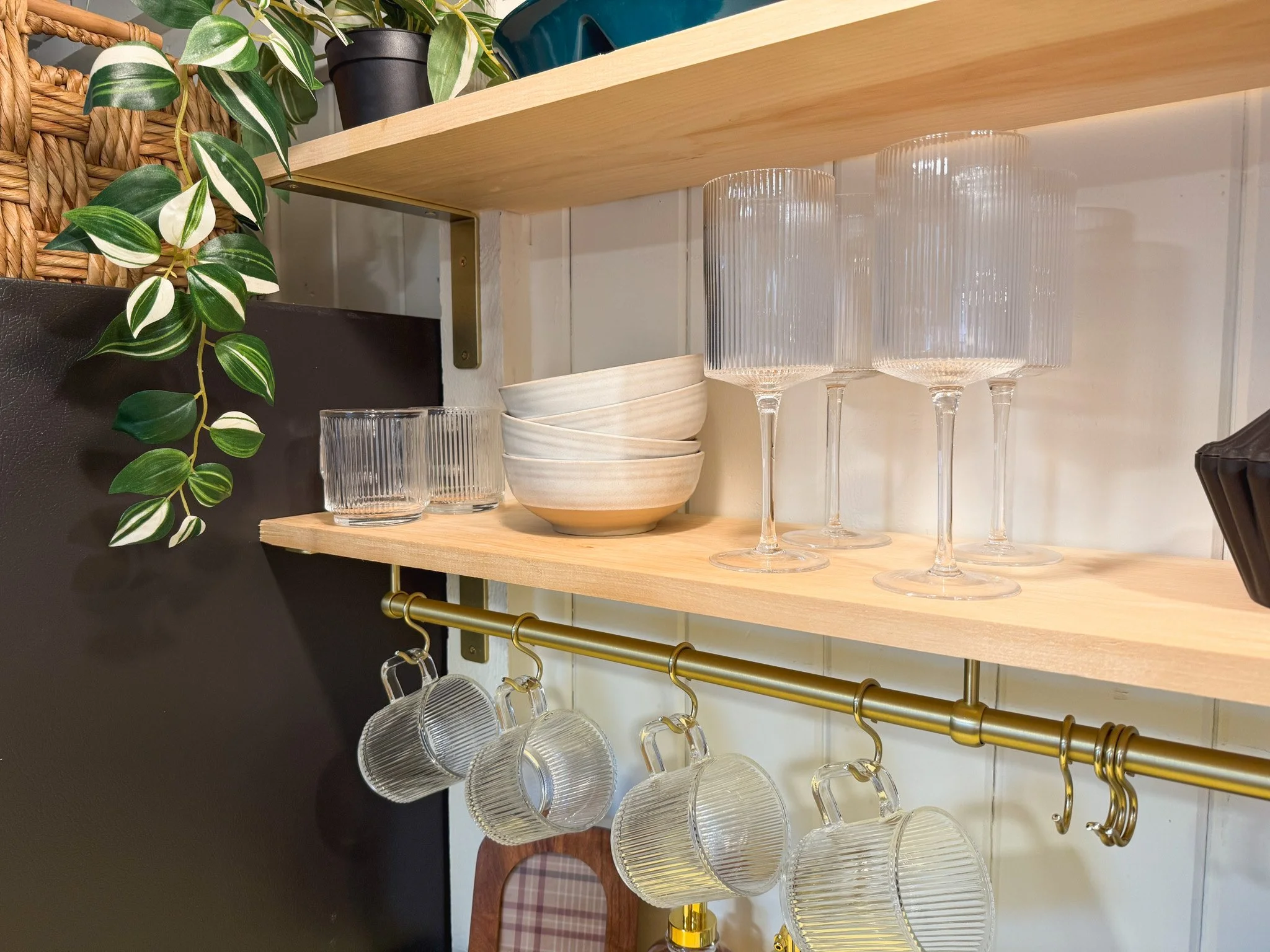 Open wooden shelf with empty drinking glasses, bowls, and wine glasses, with hanging coffee mugs below, decorated with green leafy plants nearby.