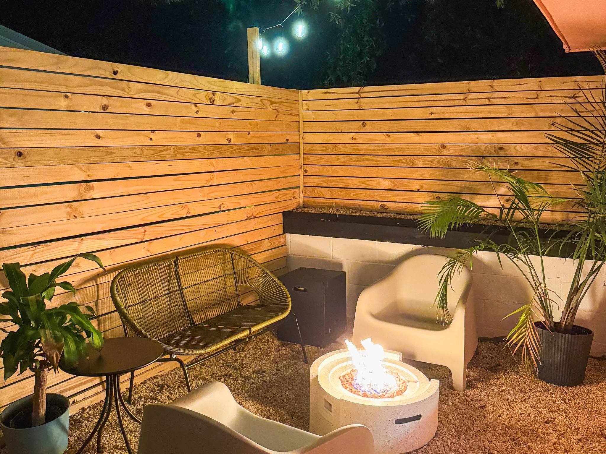 A cozy outdoor patio at night with a small fire pit, white and black chairs, a wooden privacy fence, and potted plants, illuminated by string lights.
