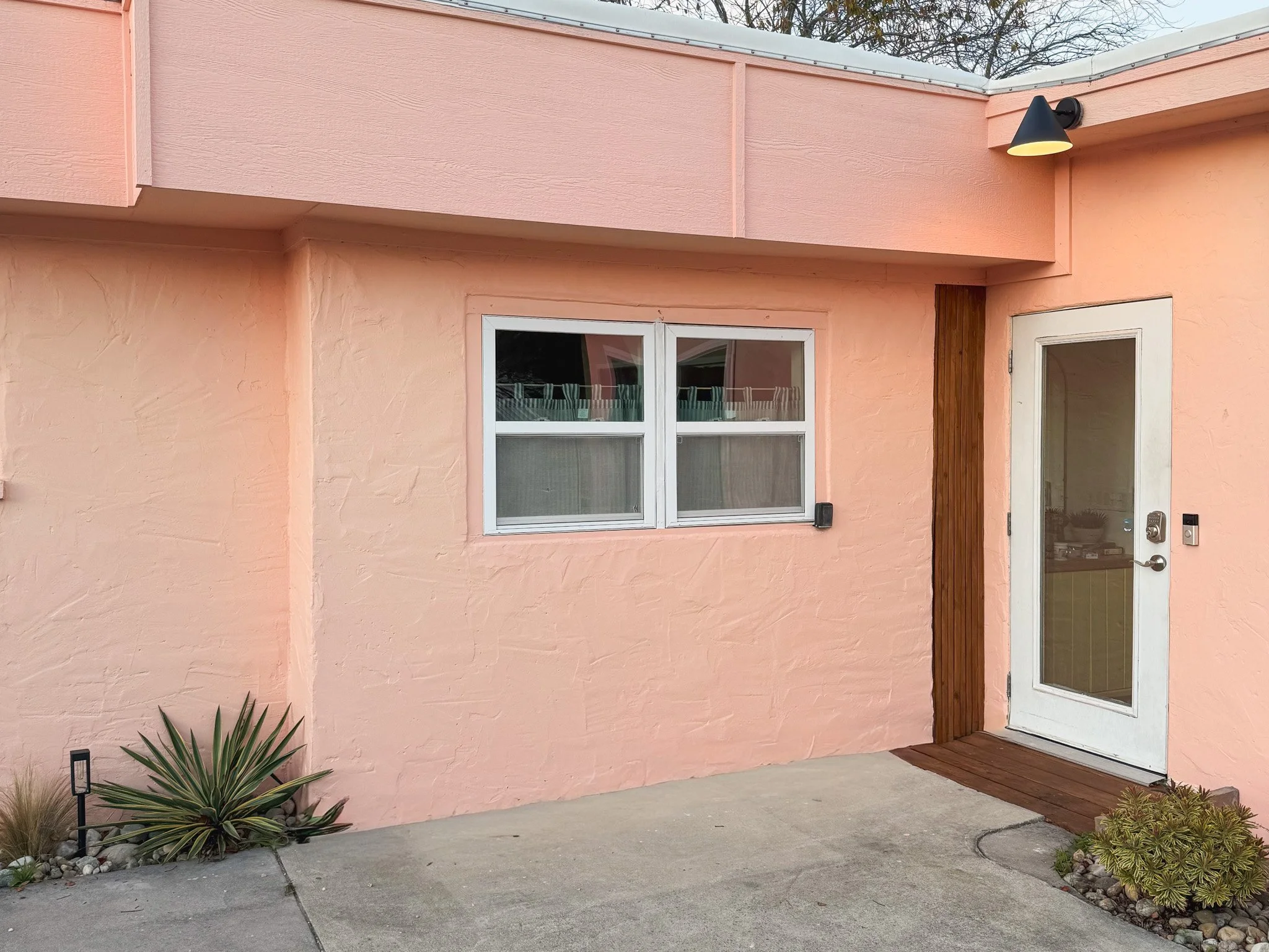 Exterior view of a pink stucco building with a white-framed window and a glass door, brown wooden accents, and desert plants near the entrance.
