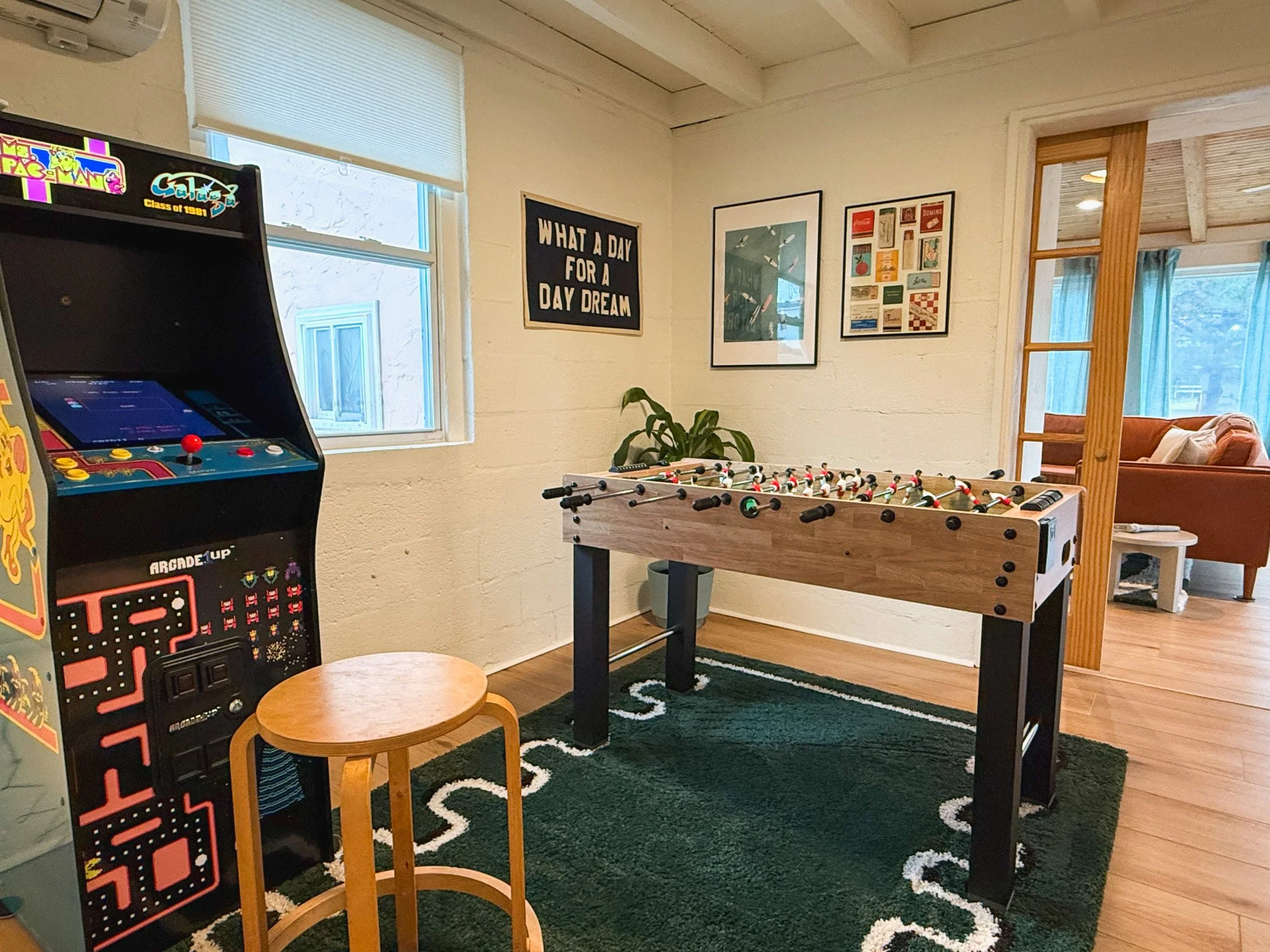 A room with a vintage arcade game on the left, a foosball table in the center, a small wooden stool in front, a black Rug with a white pattern, a window with blinds, and framed art on the wall. A sliding wooden door leads to a living area with a sofa
