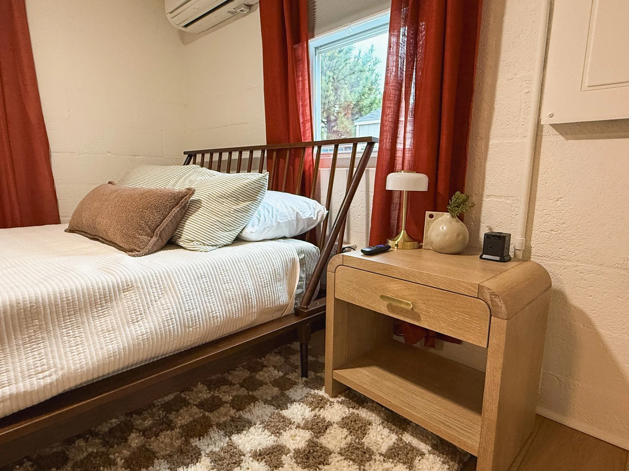 A bedroom with a bed, pillows, a window with red curtains, a nightstand with a lamp, a small clock, and a decorative vase with greenery.