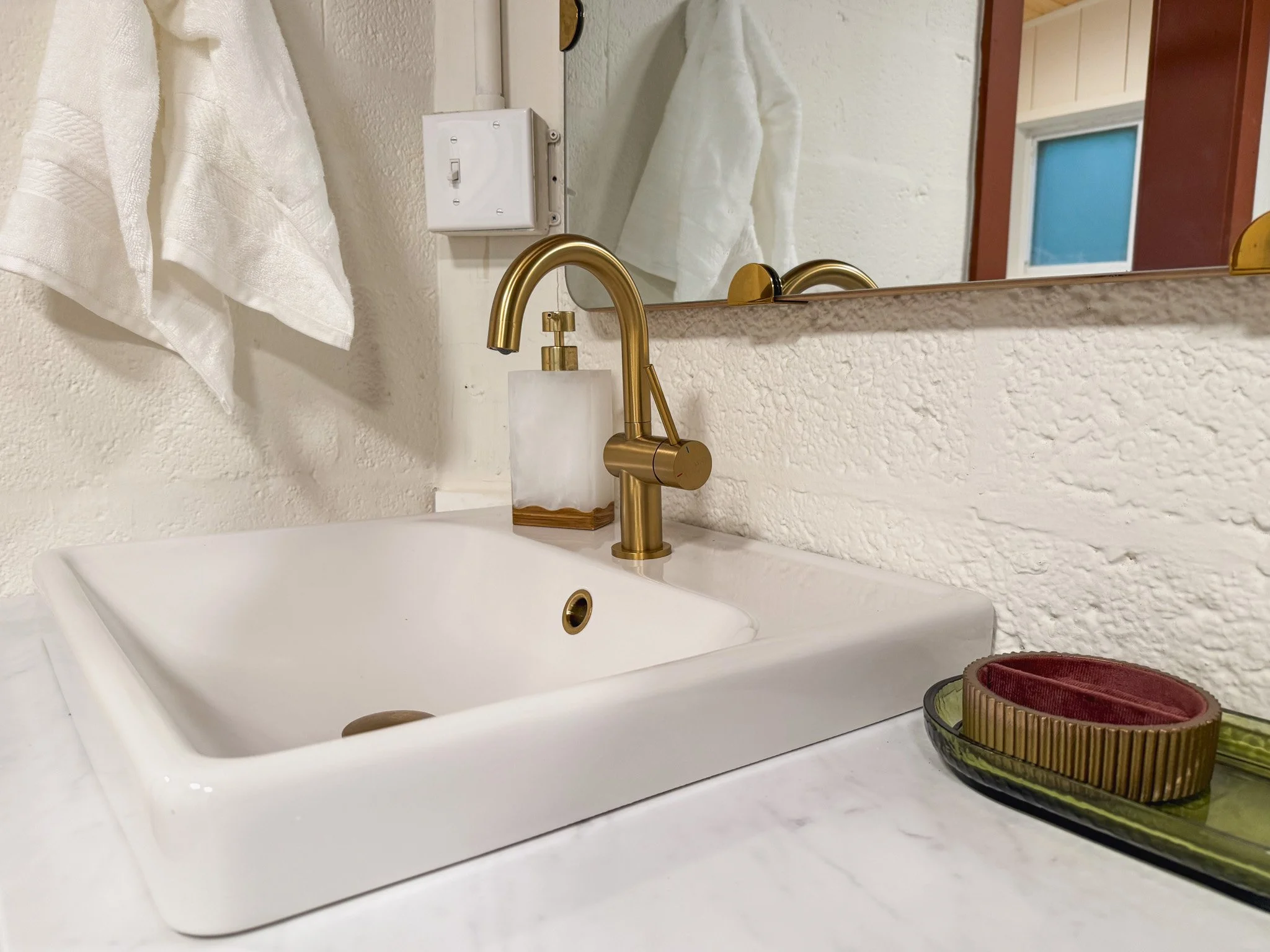 Bathroom sink with a gold faucet, white textured wall, mirror, white towel, soap dispenser, and a tray with rolled towels.