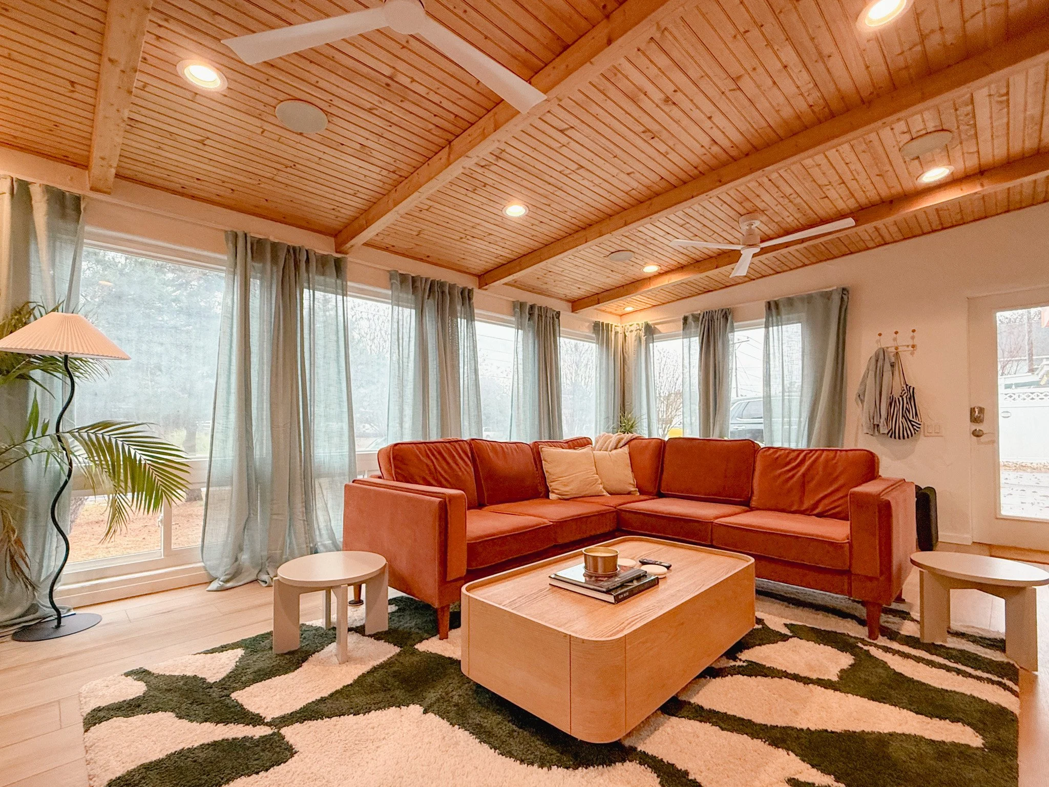 Living room with large windows, wooden ceiling, orange sectional sofa, black, white, and green patterned rug, potted plant, side tables, coffee table, and curtains.