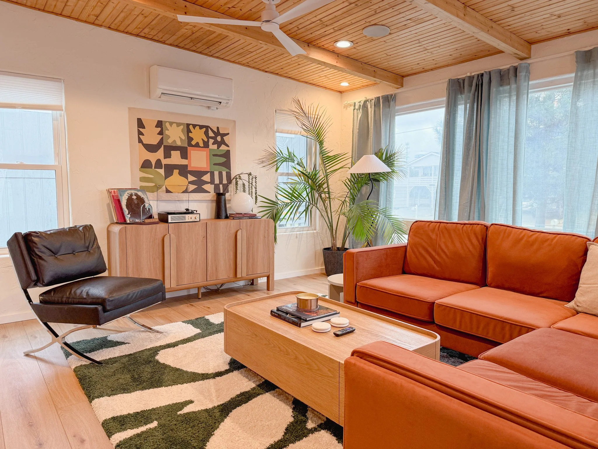 Living room with orange sectional sofa, black leather chair, wooden coffee table, large window with curtains, potted plant, floor lamp, wall art, and wooden ceiling.