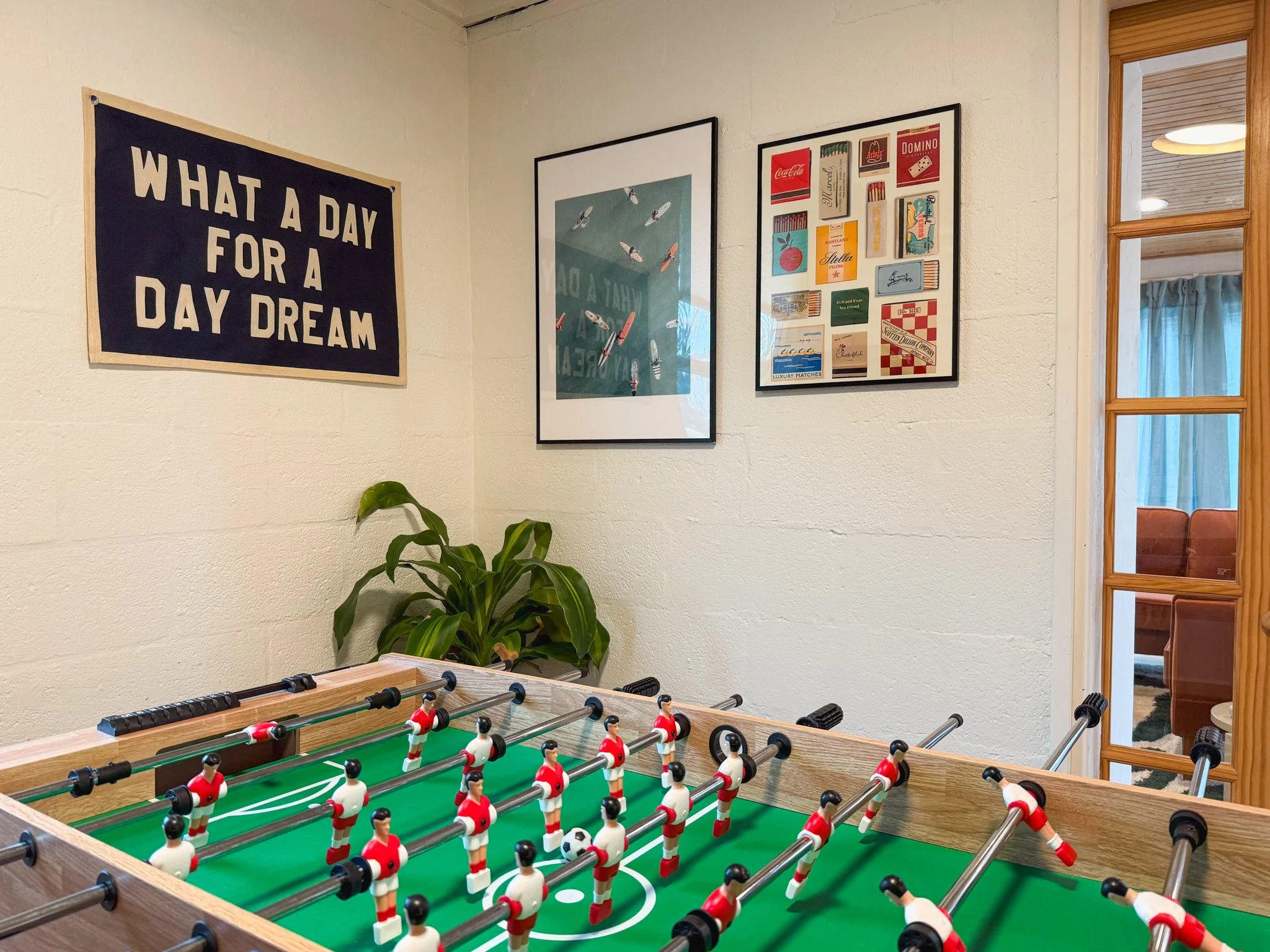 Foosball table with red and white players in a room with wall art, including a sign reading 'What a day for a daydream,' a framed picture of rockets, and a collage of matchbook covers; a potted plant is in the corner.