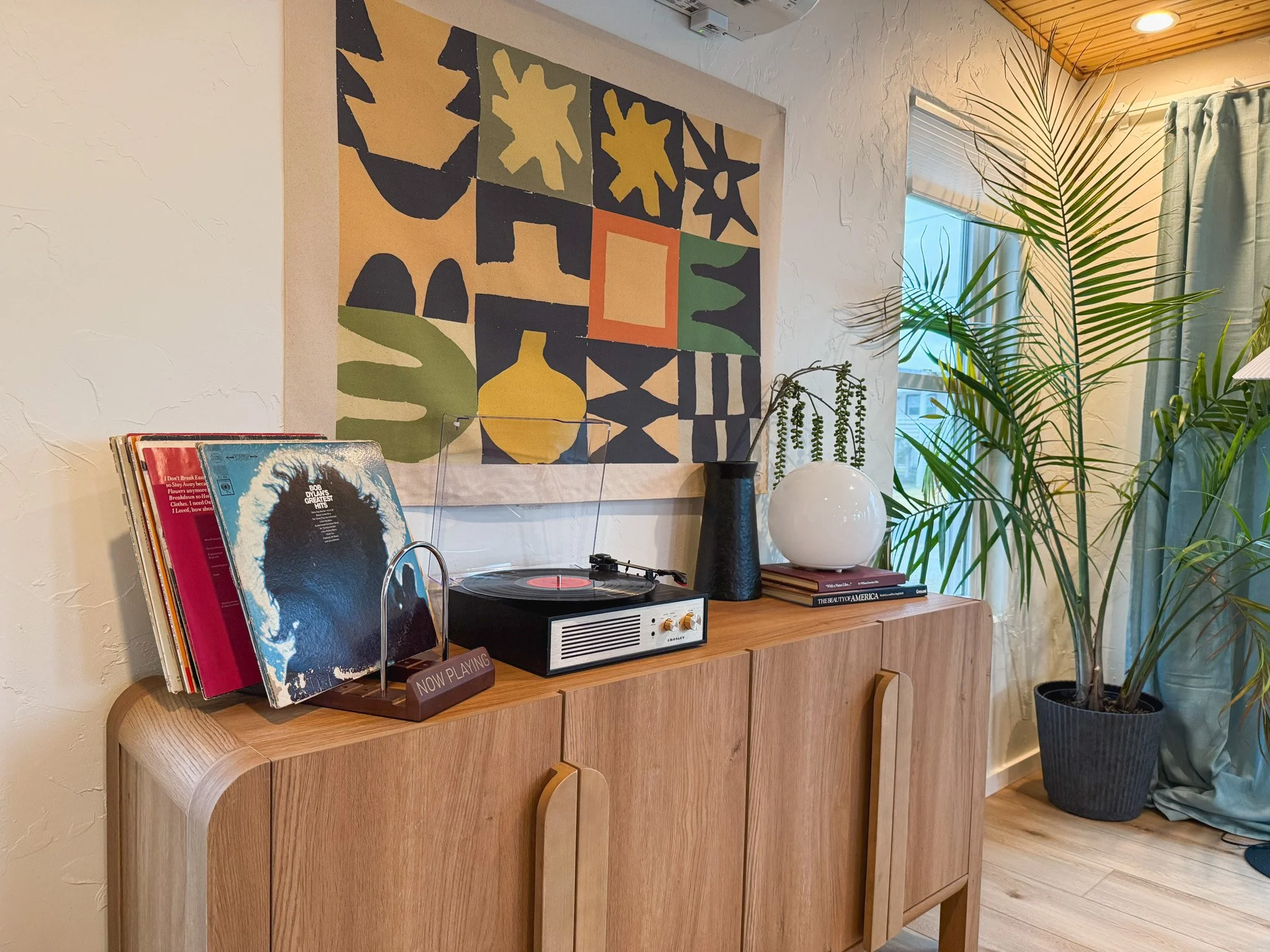 Living room decor with a wooden sideboard holding vinyl records, a record player, a small sign that says 'NOW PLAYING,' vases, and a stack of books. There is a large leafy plant in a pot, window with curtains, and abstract wall art above the sideboar
