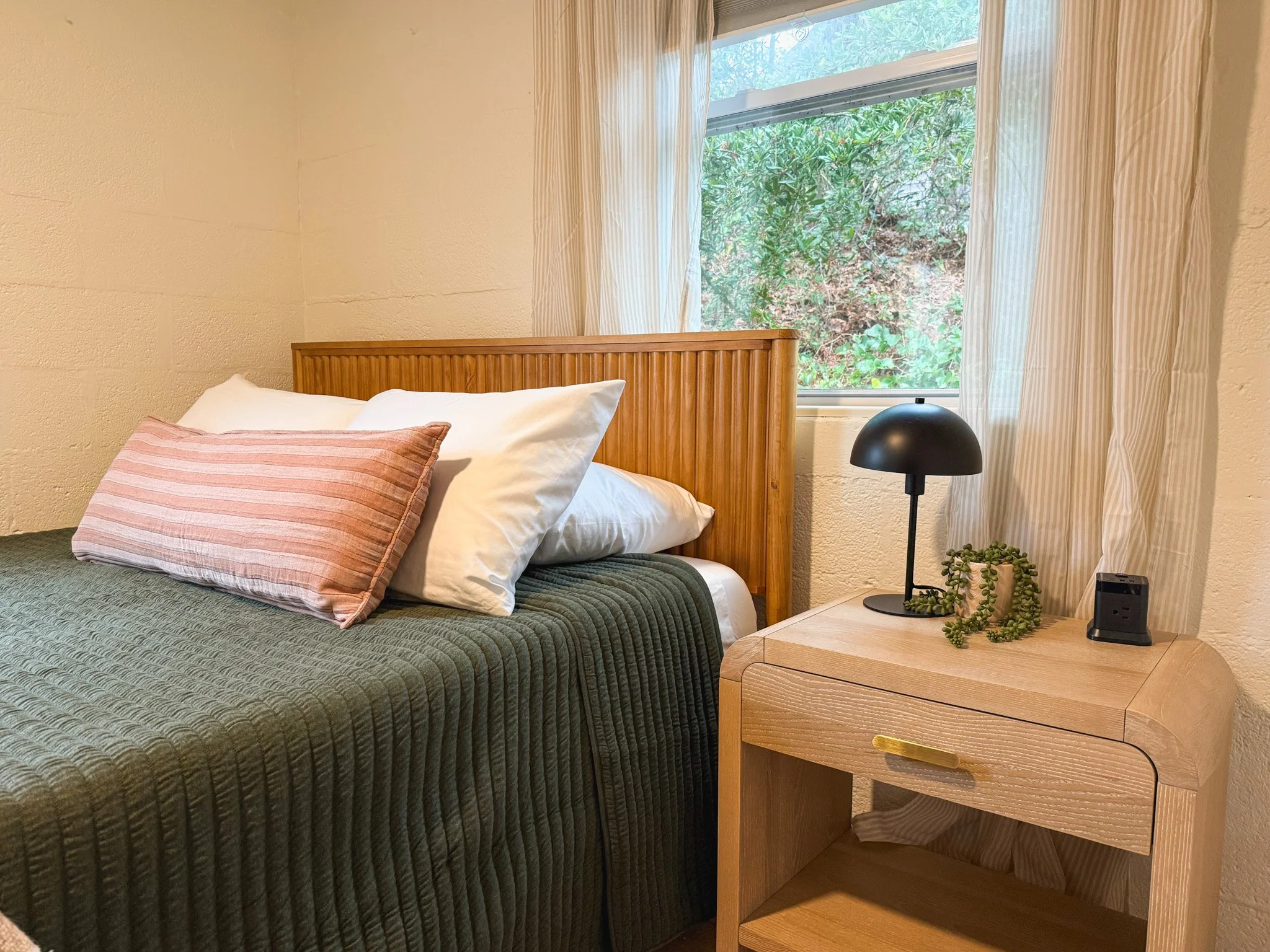 A bedroom featuring a bed with white pillows and a pink striped pillow, a wooden headboard, and a bedside table with a black table lamp, a small potted plant, and a device. There is a window with cream-colored curtains, showing greenery outside.