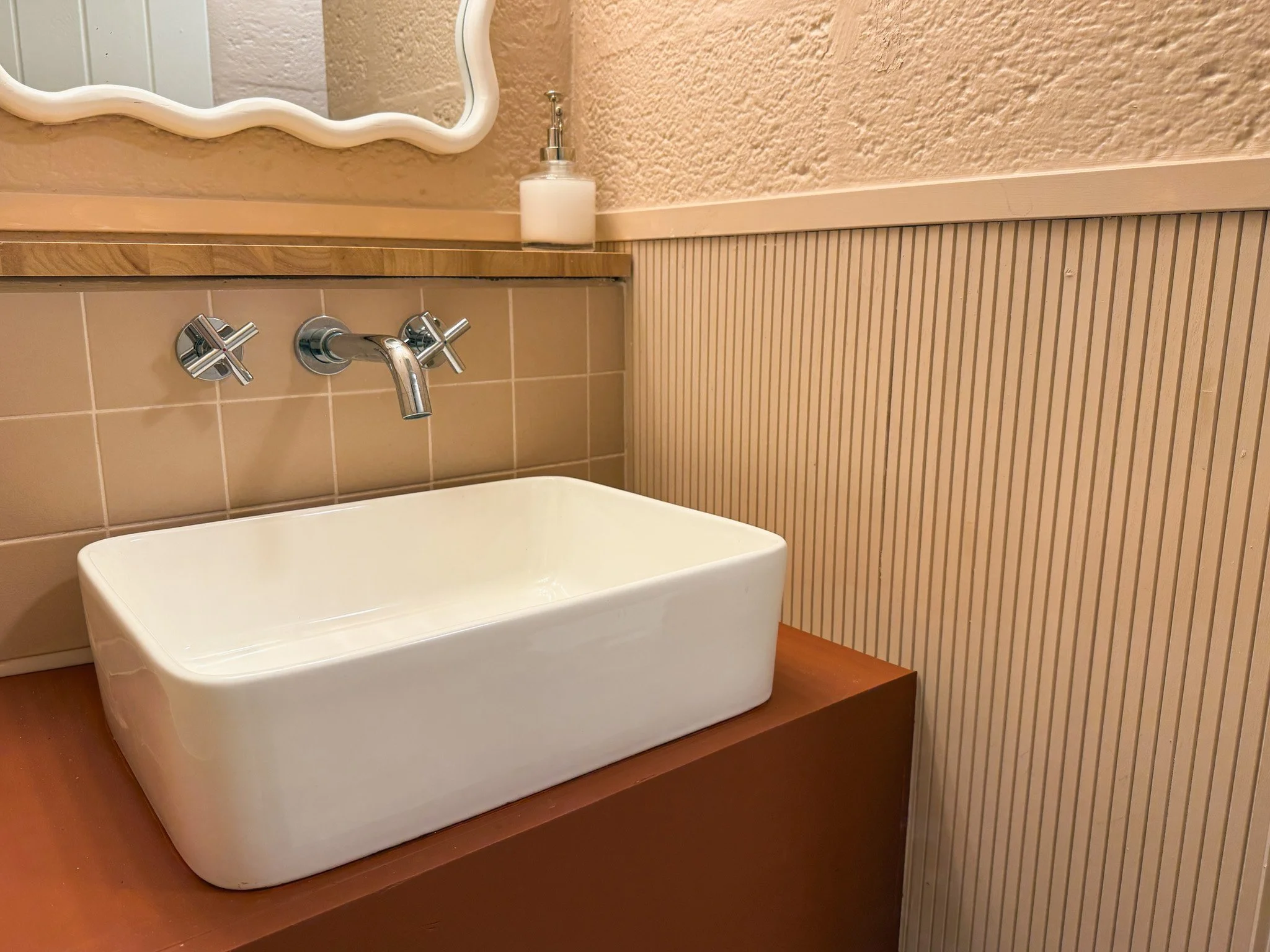 A white ceramic sink on a wooden countertop with a wall-mounted faucet and cross handles, above beige tiled wall and textured wall paneling, with a soap dispenser and a mirror above.