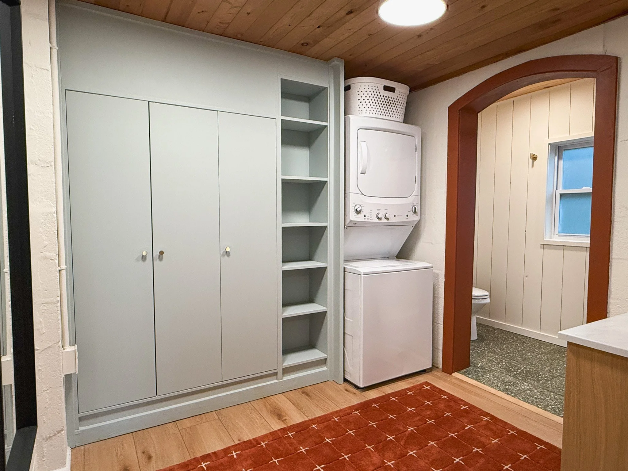 A laundry room with a white cabinet, open shelves, and a stackable washer and dryer. A basket is on top of the dryer. There is a small window and a doorway leading to a bathroom with a toilet. A red rug with a white cross pattern is on the wooden flo