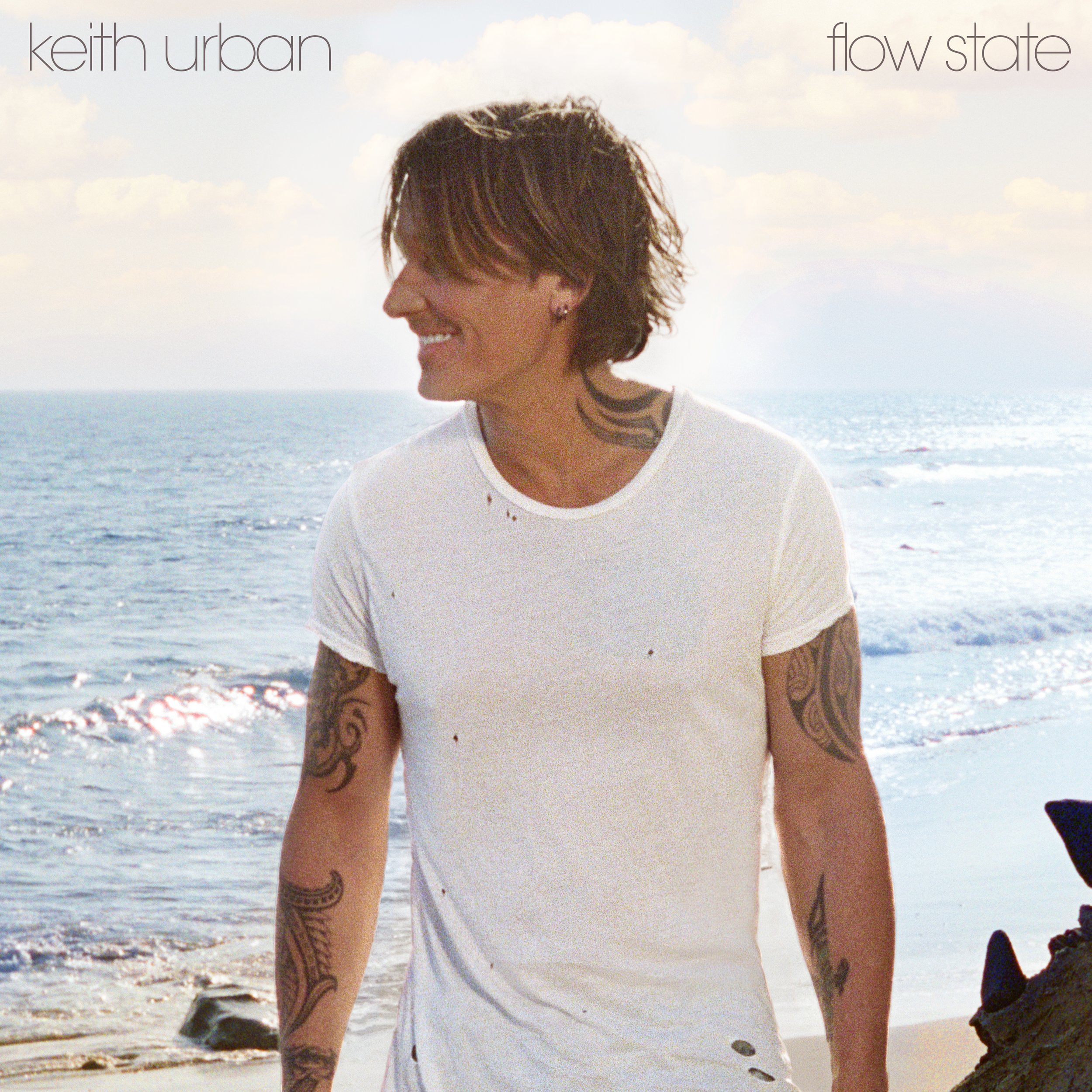 A woman with short brown hair, tattoos on her arms and neck, smiling and looking to the side, standing outdoors on a beach with ocean and sky in the background.