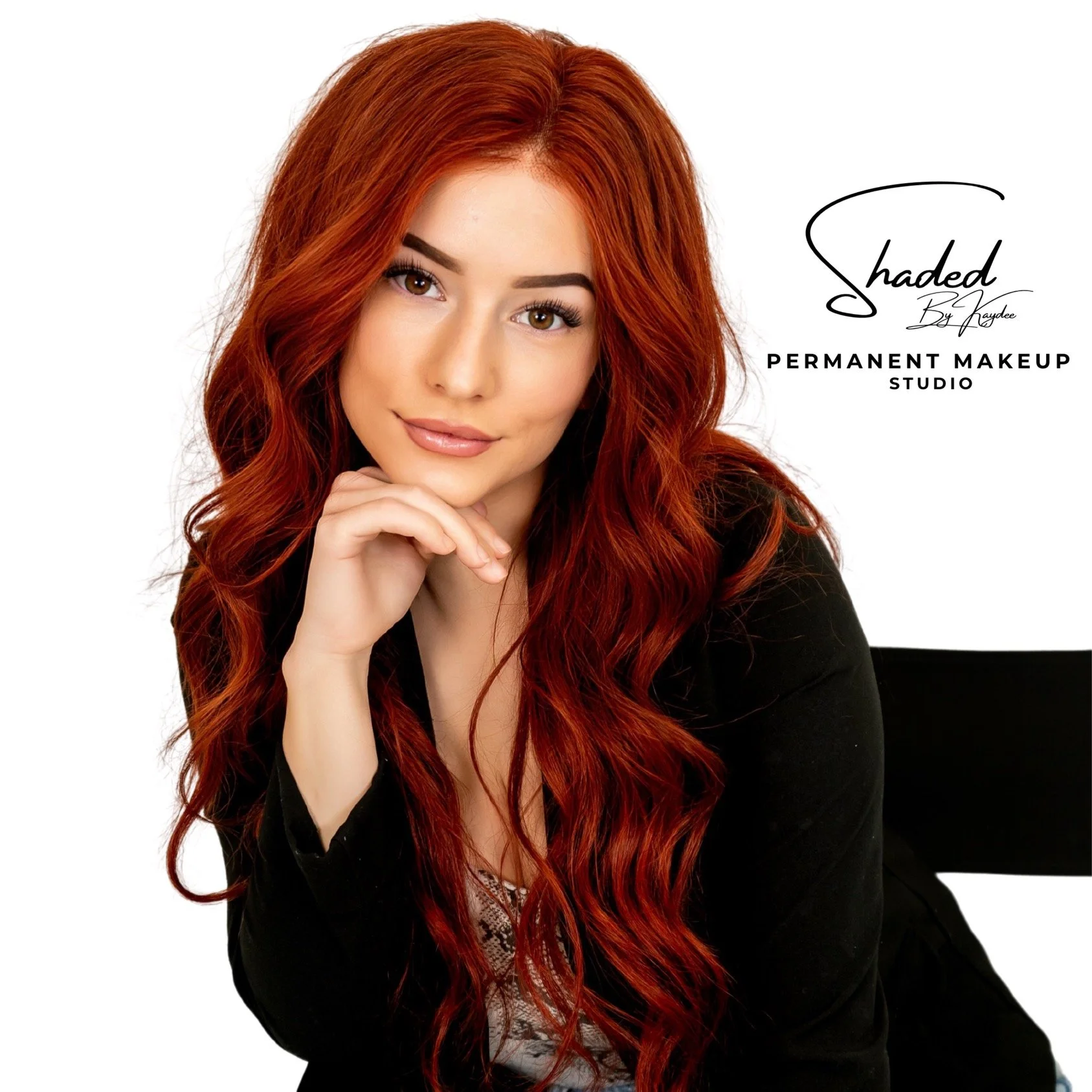A woman with long, wavy red hair, wearing makeup, poses with her chin resting on her hand, smiling gently at the camera against a white background. Text on the image reads "Shaded by Kaydee Permanent Makeup Studio."
