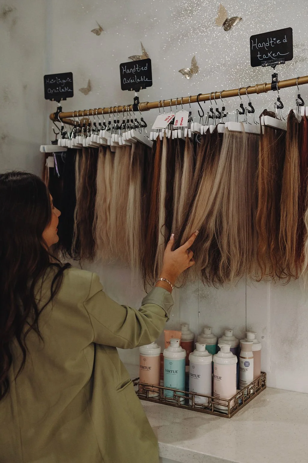 A woman looking at a display of mixed hair extensions hanging on hooks, with signs indicating availability and mentioning they are hand-tied and taken.