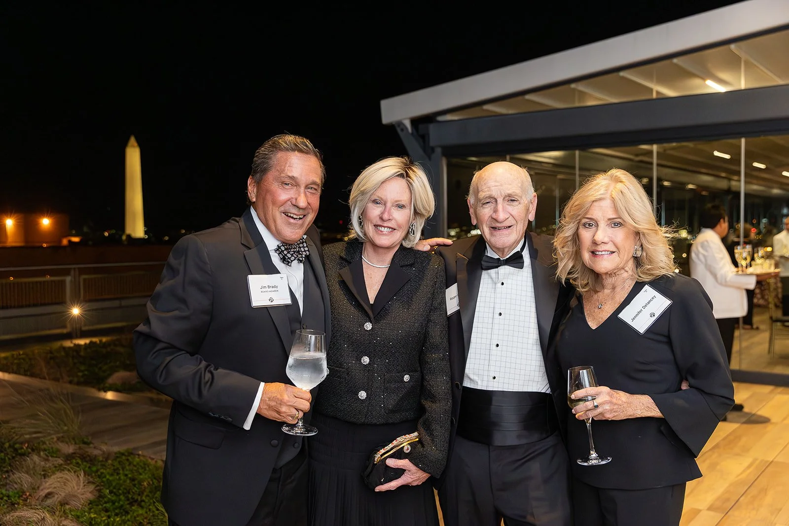 In the evening, four well-dressed friends pose with smiles and champagne glasses on the roof of the International Spy Museum with the Washington Monument visible in the background.
