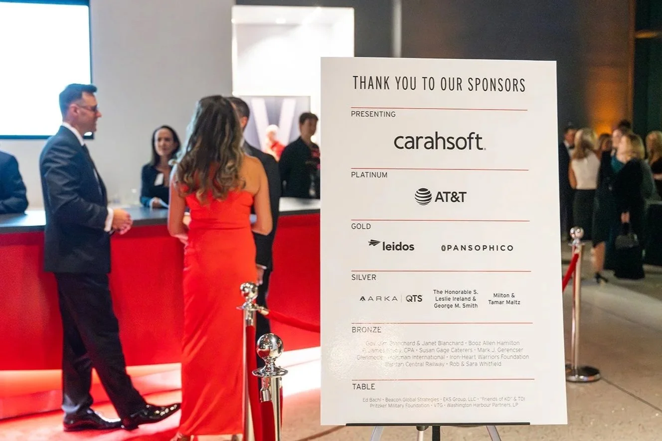 Guests arrive and interact with staff in the background as a sign in the foreground thanks sponsors for their contributions to the event.