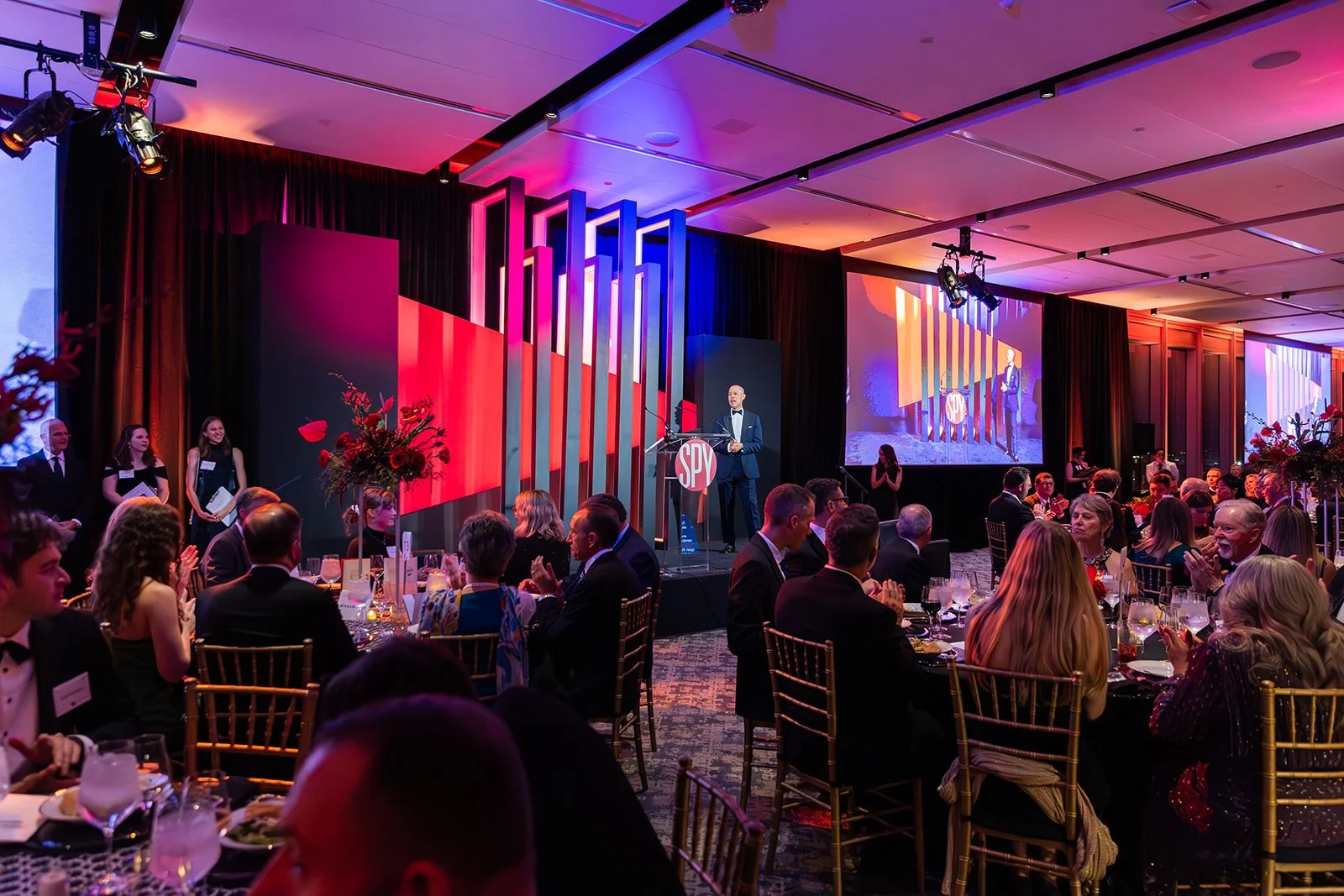 Groups of well-dressed people are sitting at round tables eating a fancy meal while Vlad Duthries speaks from the stage at the Spy Museum.