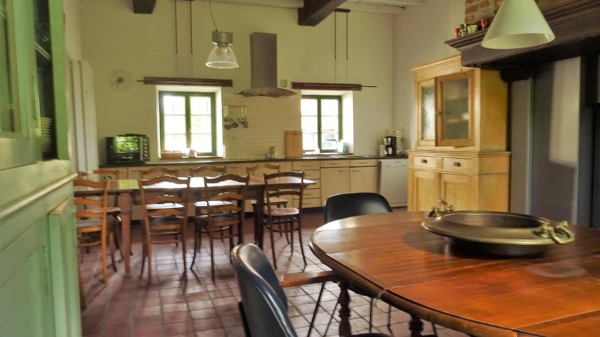 Een keuken met houten kasten, twee ramen, een eettafel met stoelen, en keukengerei. Beige muren en een stenen vloer.
