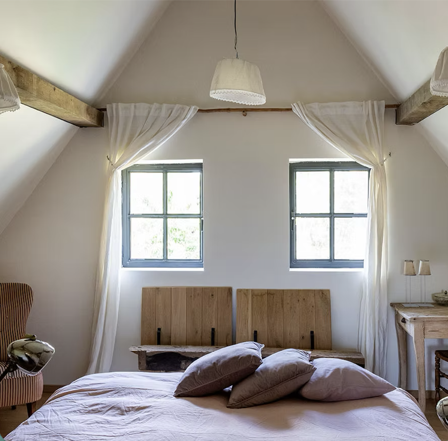 Slaapkamer met twee ramen, wit plafond en gordijnen, houten bedankers, bed met roze dekbedovertrek en kussens, houten nachtkastje en lamp, houten beugel op muur.