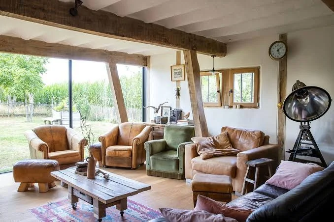 Een gezellige woonkamer met grote ramen, banken en fauteuils van verschillende kleuren, een houten salontafel, planten en decoraties, met uitzicht op een groene tuin.