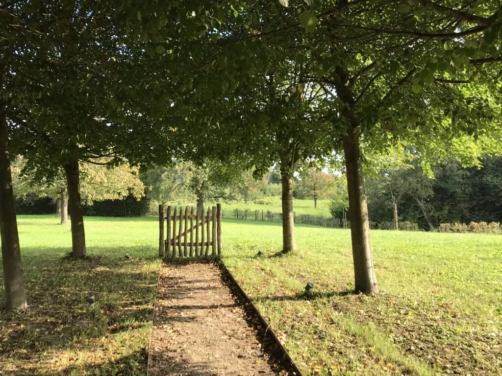 Een pad met een kleine houten poort tussen drie bomen, leidt naar een boomgaard achter vakantiewoning De Waterkriek