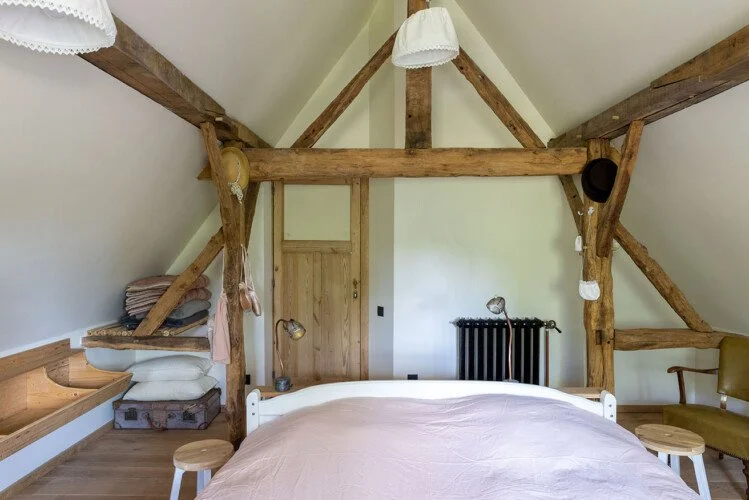 Een slaapkamer met houten balken en een groot bed, daarnaast een old-fashioned radiator en een vintage stoel in de hoek.