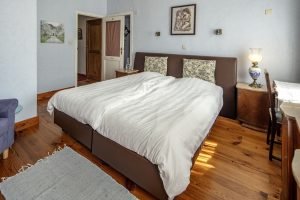 Een slaapkamer met een groot tweepersoonsbed, witte lakens, een nachtkastje met een lamp, en muren versierd met schilderijen.